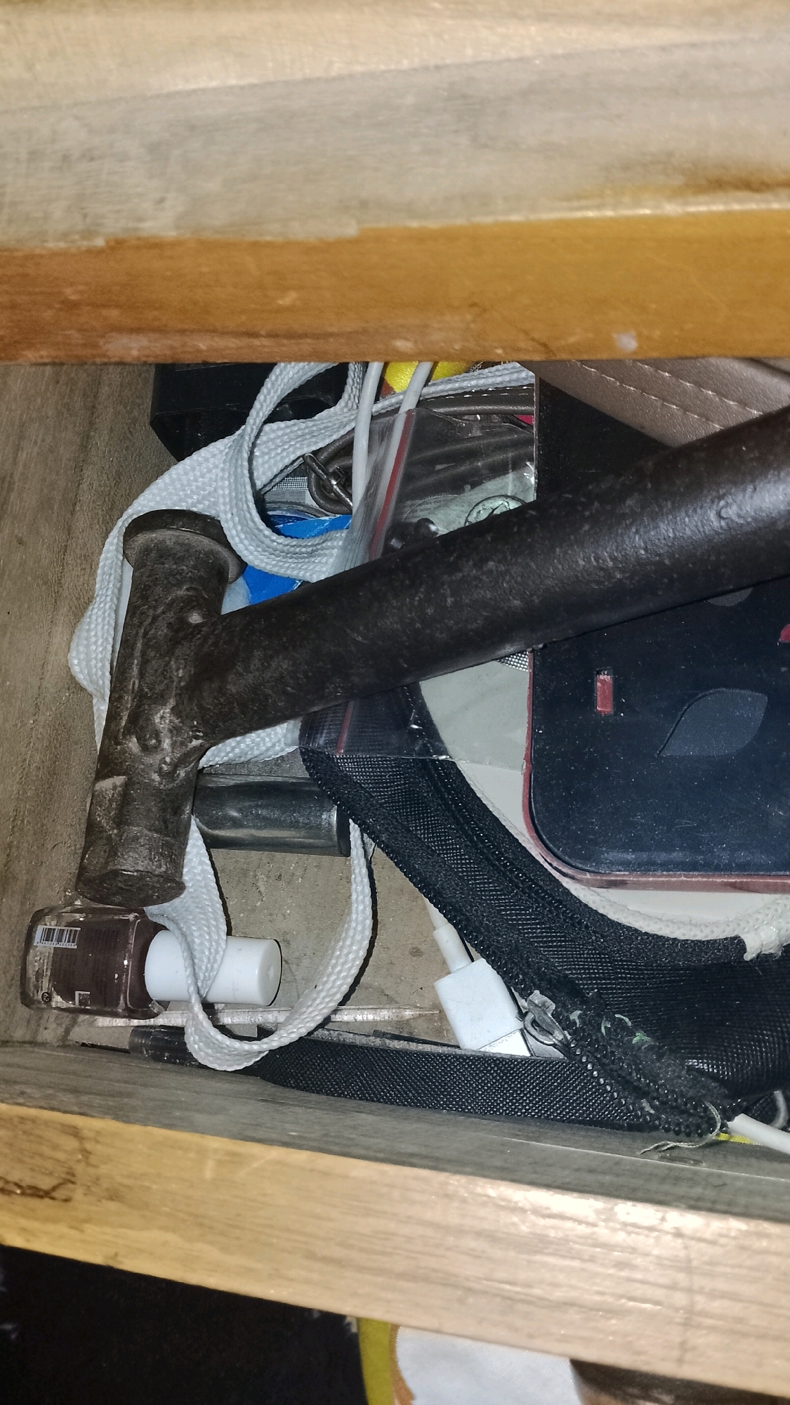 The image shows a cluttered drawer with various items. In the foreground, a dark metal hammerhead rests on a white strap. To the left of the hammerhead is a bottle of brown nail polish. Behind the hammer, a black and red electronic device is visible, partially covered by a black mesh bag. Various wires and other unidentifiable objects are scattered throughout the drawer. The drawer appears to be made of wood. The scene is dimly lit, suggesting an indoor environment. The image does not provide any information about the location in the city of Mulyo Haji, Indonesia.
