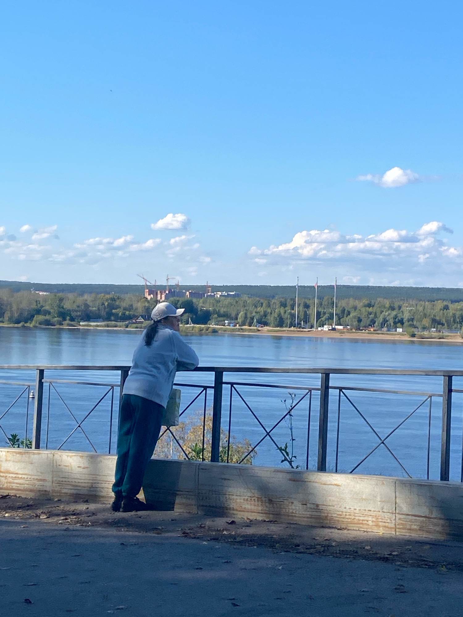 The image shows a person wearing a cap, light-colored jacket, and dark pants, leaning on a metal railing and looking out over a wide body of water. Behind the water, a line of trees forms a distant horizon, with some buildings and construction cranes visible in the background. The sky is blue with scattered white clouds. This scene appears to be from the embankment along the Kama River in the city of Perm, Russia, likely near a viewpoint or park area.