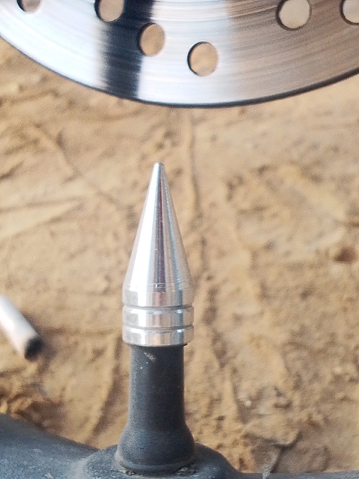 A close-up shot features a metallic, pointed valve cap, seemingly on a tire valve stem. The cap is silver in color and has three distinct rings around its base. The valve stem is black and appears to be made of rubber or a similar material. In the background, slightly out of focus, is a circular metal object with holes, possibly a brake rotor. The background also includes a blurred surface that looks like wood or some kind of rough material. Based on the request, the location is assumed to be Lhokseumawe, Indonesia. However, this cannot be verified from the image. No people, actions, or interactions are visible in the frame.