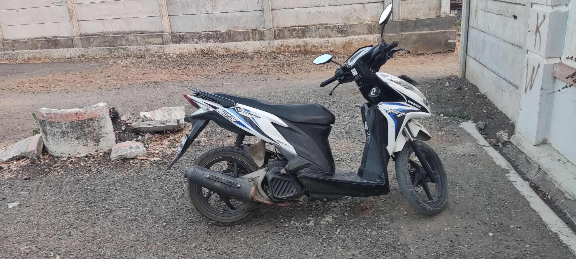 A white and black Honda motorcycle is parked on a gravelly surface next to a concrete wall in Pakusamben, Indonesia. The motorcycle is a scooter-style model, with the front wheel turned slightly to the left. It has a black seat, black handlebars, and silver mirrors. The body of the motorcycle is predominantly white with blue and black accents. There is a white concrete wall with some graffiti on it in the background, and a broken concrete pipe lying on the ground to the left of the motorcycle. The ground is covered in loose gravel and dirt.