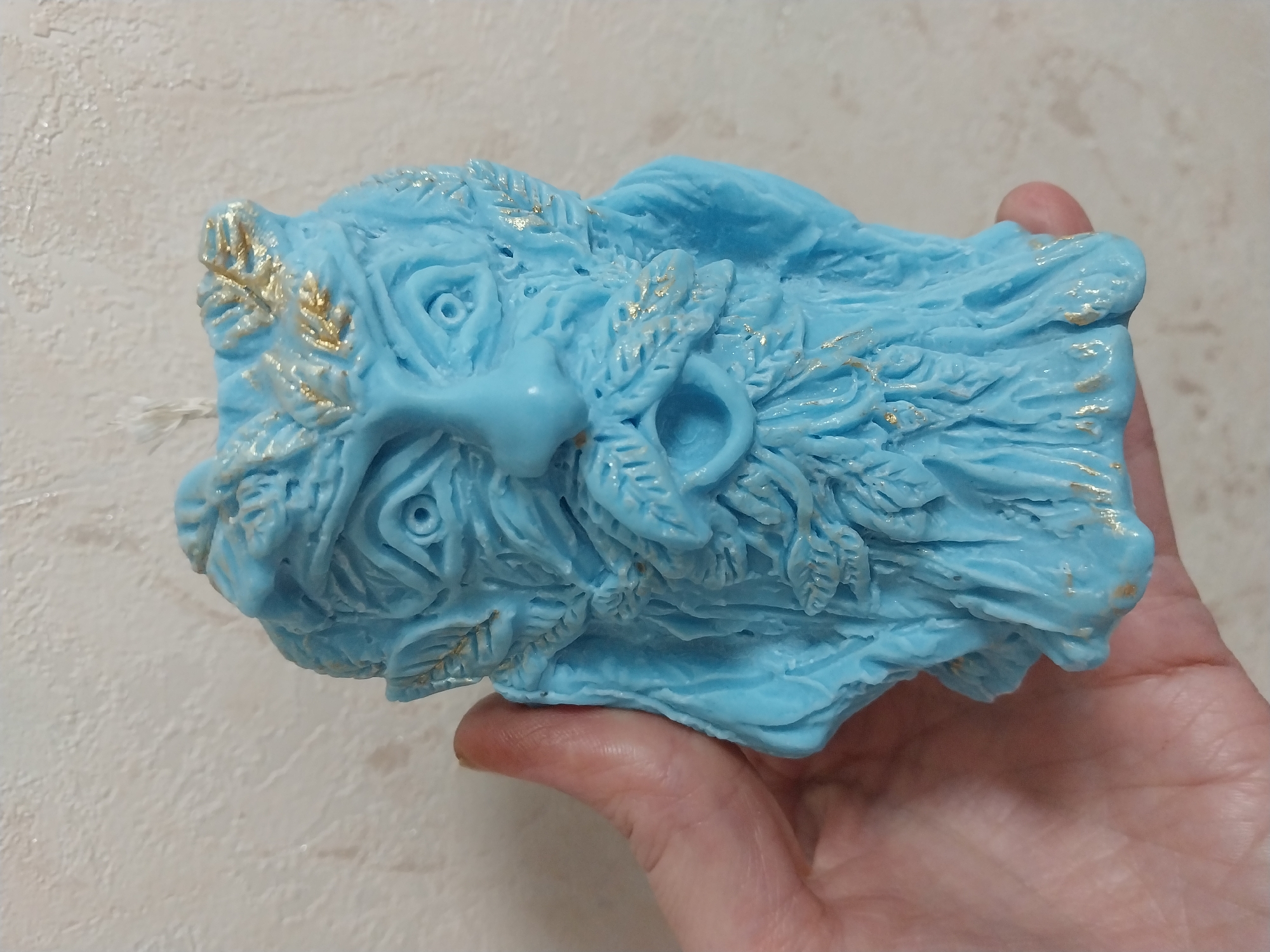 A light blue, intricately sculpted candle in the shape of a mythical face, possibly a nature spirit or deity, is held in a person's hand against a textured, light-colored wall. The candle features a detailed face with wide eyes, a prominent nose, and a flowing beard adorned with leaf-like patterns. Gold accents highlight some of the sculpted details, such as the leaves and hair. The hand holding the candle is positioned on the left side of the frame, obscuring part of the candle's body. The background wall appears to have a slightly rough, plaster-like texture. There is no indication of the city of Dnipro, Ukraine, or any other specific location.
