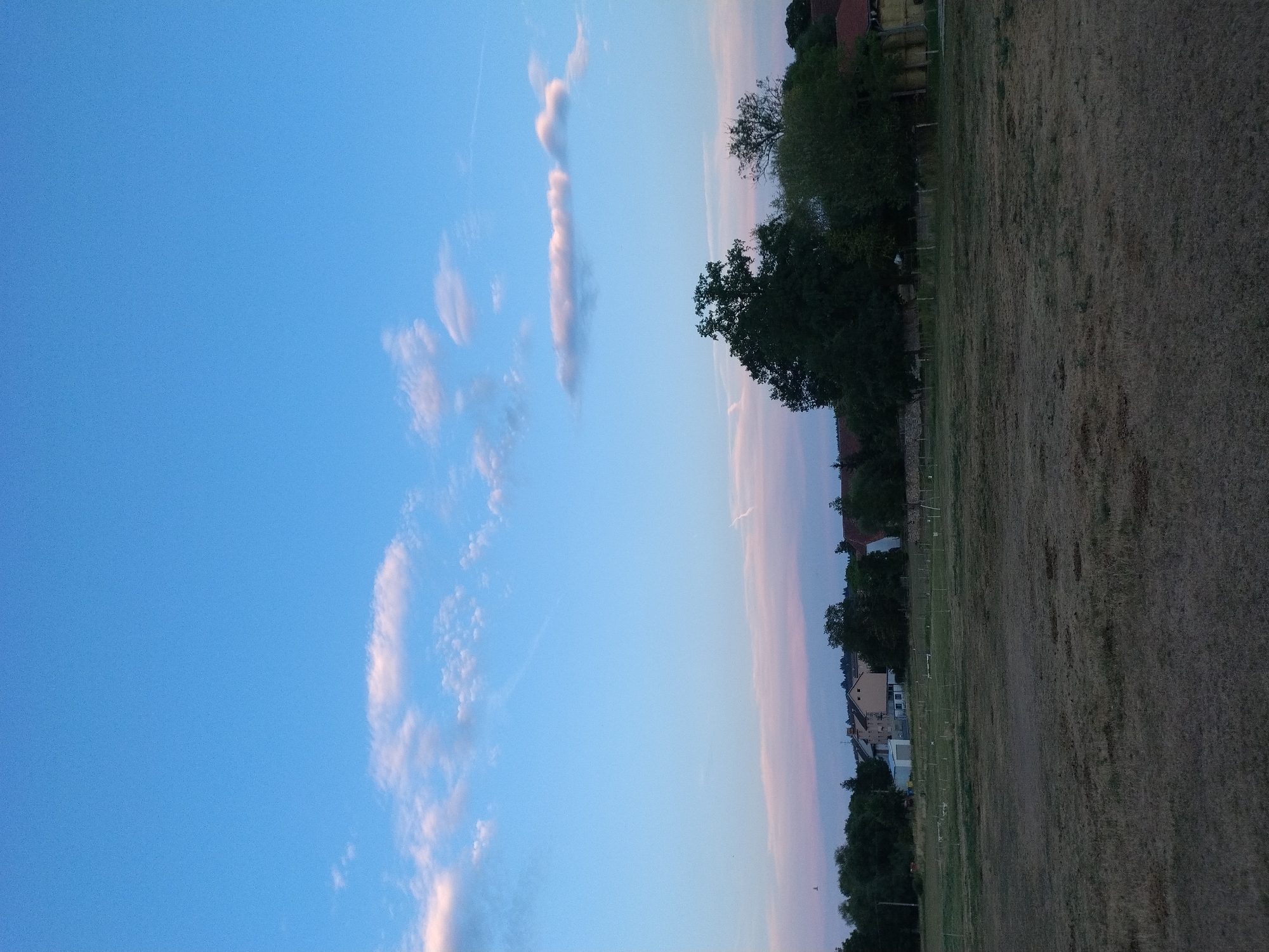 The image shows a rural scene in Dobev, Czechia, at dusk. The sky is a gradient of blue, with streaks of pink and white clouds near the horizon. In the middle ground, a line of trees and buildings forms the edge of a village. The buildings appear to be residential, with pitched roofs and varying architectural styles. Some are light-colored, while others are darker. A large, dark green tree dominates the right side of the frame, casting a shadow. In the foreground, a dry, grassy field stretches across the bottom of the image, with sparse vegetation and some faint markings that suggest a fence or boundary. The overall impression is one of peaceful, late-day rural tranquility.