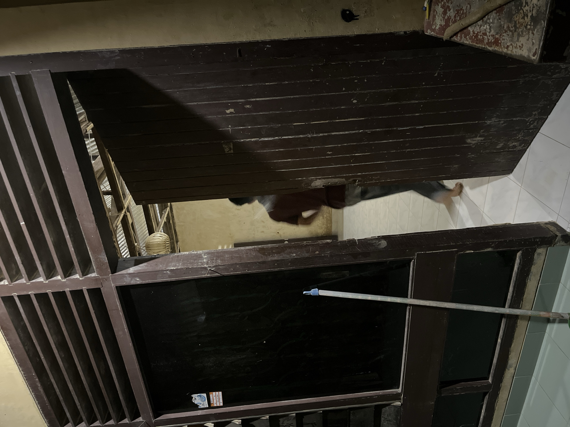 The image shows a person walking in what appears to be an indoor setting, possibly a hallway or a covered walkway, judging by the tiled floor and the wooden structures. The lighting suggests it might be evening or indoors, with artificial light sources.
The person is wearing dark clothing and is captured mid-stride. Their body is angled away from the camera, and their legs are visible. The background shows wooden elements, possibly part of a doorway or window frame, painted a dark brown color. There are also some glimpses of what could be other parts of a building, including a lighter-colored wall and some ceiling structures.
A prominent object in the foreground is a long, metallic pole, which appears to be a broom or a mop handle, positioned horizontally across a dark, reflective surface, likely a window or a glass panel. The pole has a light blue cap on one end and a metallic end on the other.
The overall scene has a slightly worn or aged aesthetic, with some of the wooden surfaces showing signs of age or wear. There is no visible text in the image that is clearly legible. The setting seems to be domestic or residential, given the nature of the interior elements. The activity depicted is simple movement, with the person walking through the space. No specific emotional cues are evident due to the distance and the pose of the person.