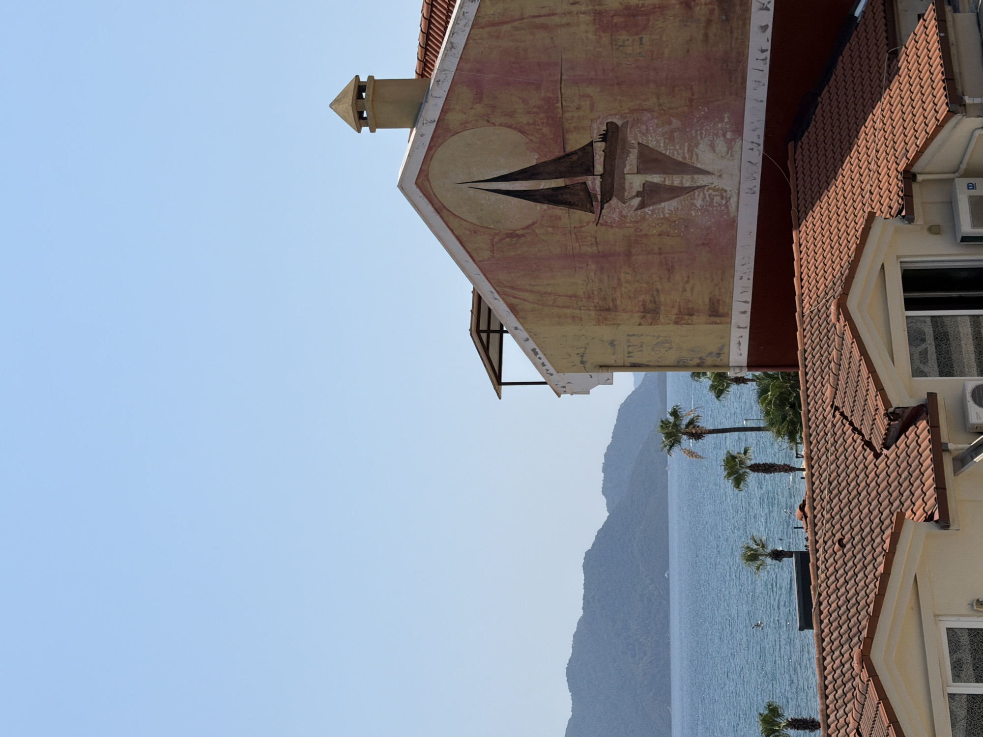 The image captures a view from a rooftop in Marmaris, Turkey, overlooking the sea and distant mountains. In the foreground, two terracotta-tiled roofs of residential buildings are visible. The building on the right features a mural of a sailboat silhouetted against a large, pale circle on its yellowish-orange facade. A chimney with a conical top extends from this building. In the middle ground, the blue sea is dotted with palm trees and a person on a paddleboard. The background consists of hazy, blue-green mountains under a clear sky.