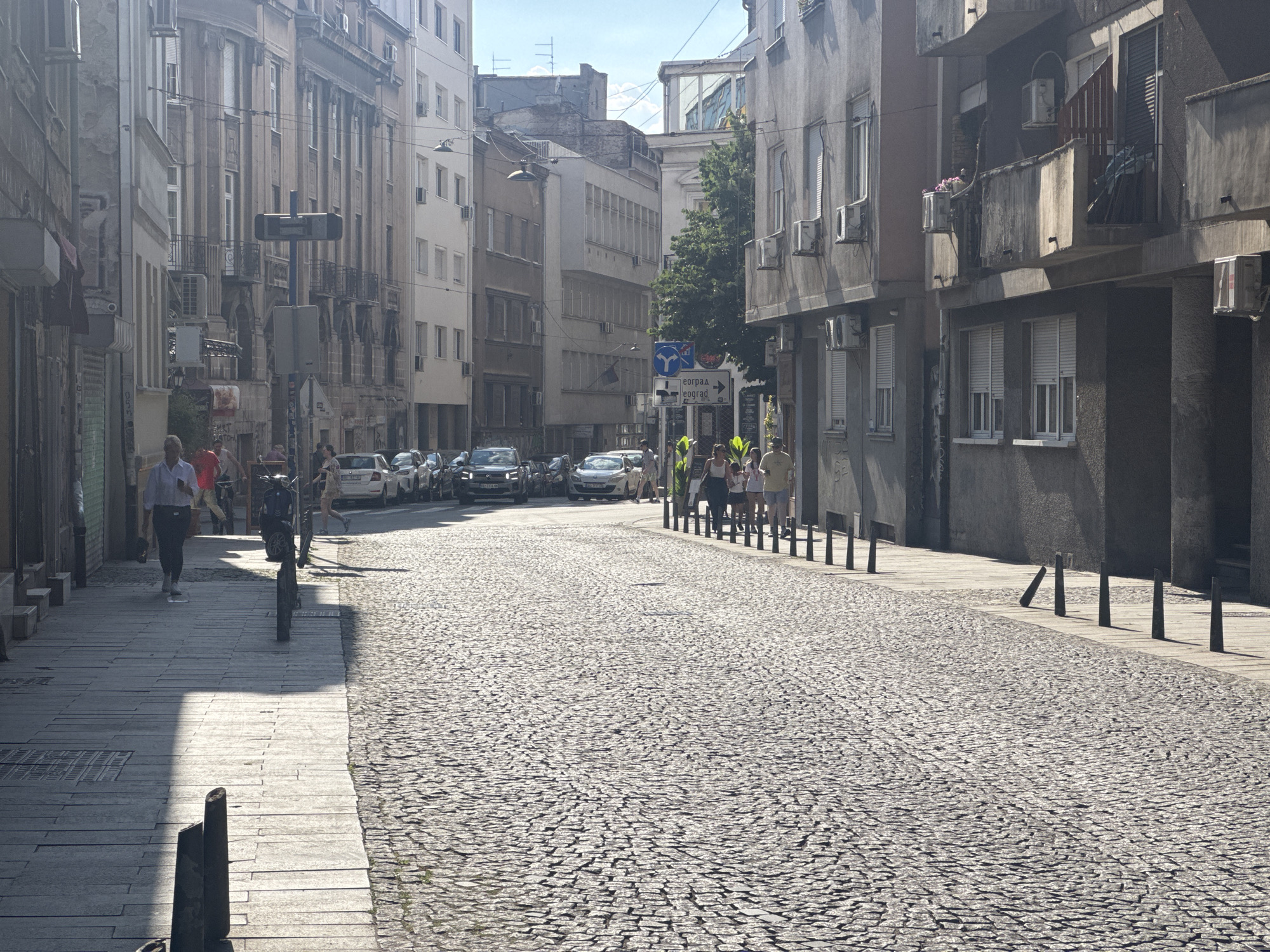 The image depicts a street scene in Belgrade, Serbia, on a sunny day. The street is paved with cobblestones, and the sidewalks are made of large stone slabs. Buildings line both sides of the street, with a mix of architectural styles. On the left, the buildings appear older, with weathered facades and balconies. On the right, the buildings are more modern, with balconies and air conditioning units visible.
Several people are present in the scene. On the left sidewalk, a man in a white shirt and dark pants is walking away from the camera. A motorcycle is parked near him. Further down the sidewalk, a group of people are visible, some walking and some standing. On the right side of the street, a group of people are standing near the sidewalk, appearing to be conversing.
Cars are parked and driving on the cobblestone street. In the distance, street signs are visible, one with Cyrillic lettering. The sky is clear and blue. The scene suggests a typical urban environment in Belgrade, with a blend of old and new architecture and daily life unfolding on the street.