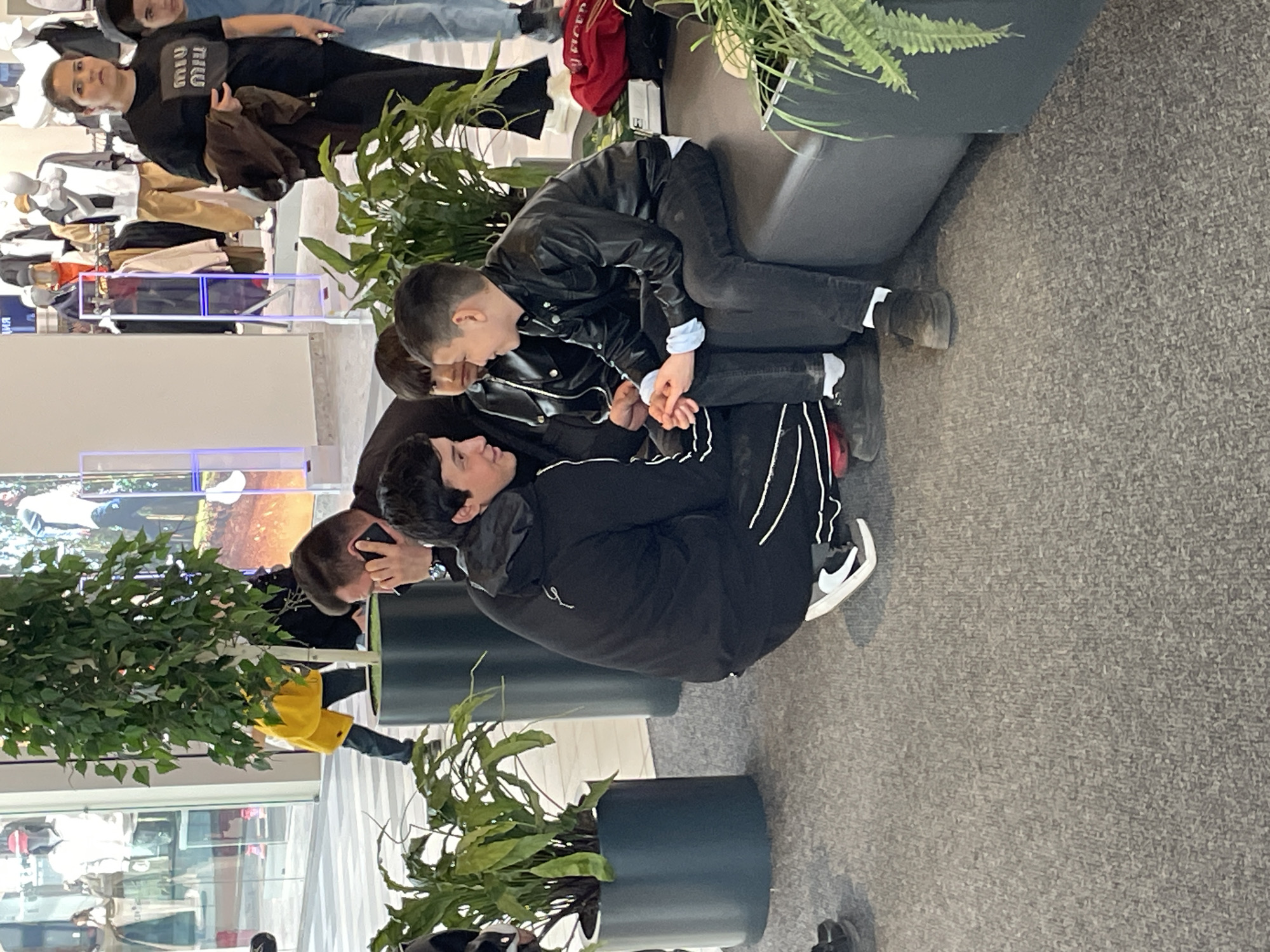In a brightly lit interior, possibly a shopping mall or commercial space in Саларьево, Russia, a group of people is gathered around seating areas adorned with potted plants. Three individuals are situated on a dark gray sofa; two are interacting closely, while another sits nearby. One person is wearing a black leather jacket and dark pants, while another wears a black hooded jacket with white stripes on the sleeves and dark pants. Shoes of varying colors are visible on their feet. The environment includes retail displays with mannequins and clothing racks in the background, as well as additional individuals. The floor is covered with a textured gray carpet.