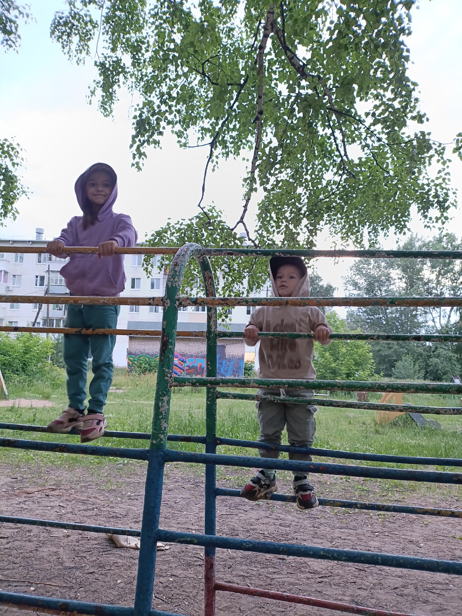 Two children, a girl and a boy, stand on a metal playground structure outdoors. The girl, wearing a light purple hooded sweatshirt and teal pants, is on the left. The boy, wearing a light brown hooded sweatshirt and grey pants, is on the right. They are both looking in different directions. The playground structure appears to be made of metal bars painted in green and yellow. In the background, there is a multi-story building, trees with green leaves, and a cloudy sky. The ground is covered with dirt and grass. The image is likely taken in Chaikovsky, Russia, as indicated by the residential building style and vegetation.