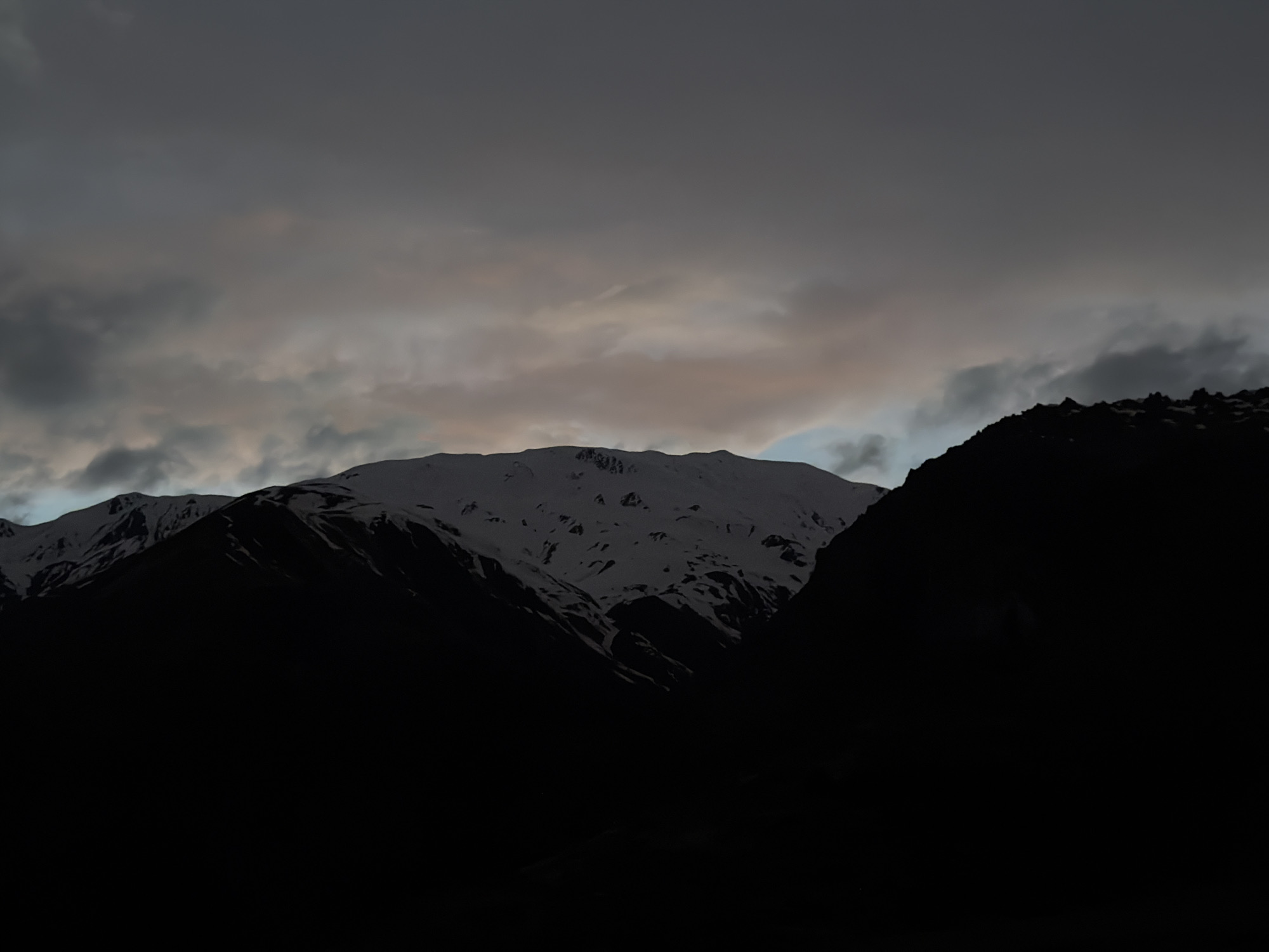 The image depicts a mountainous landscape, likely near Pkhelshe, Georgia, under an overcast sky. The foreground is dominated by dark, silhouetted mountain ranges, obscuring the terrain. A mid-range mountain peak is partially covered in snow, with dark crevices and shadows revealing its rugged texture. The sky is filled with gray and white clouds, with some hints of lighter tones suggesting a break in the cloud cover. The overall scene conveys a sense of remoteness and dramatic weather conditions.