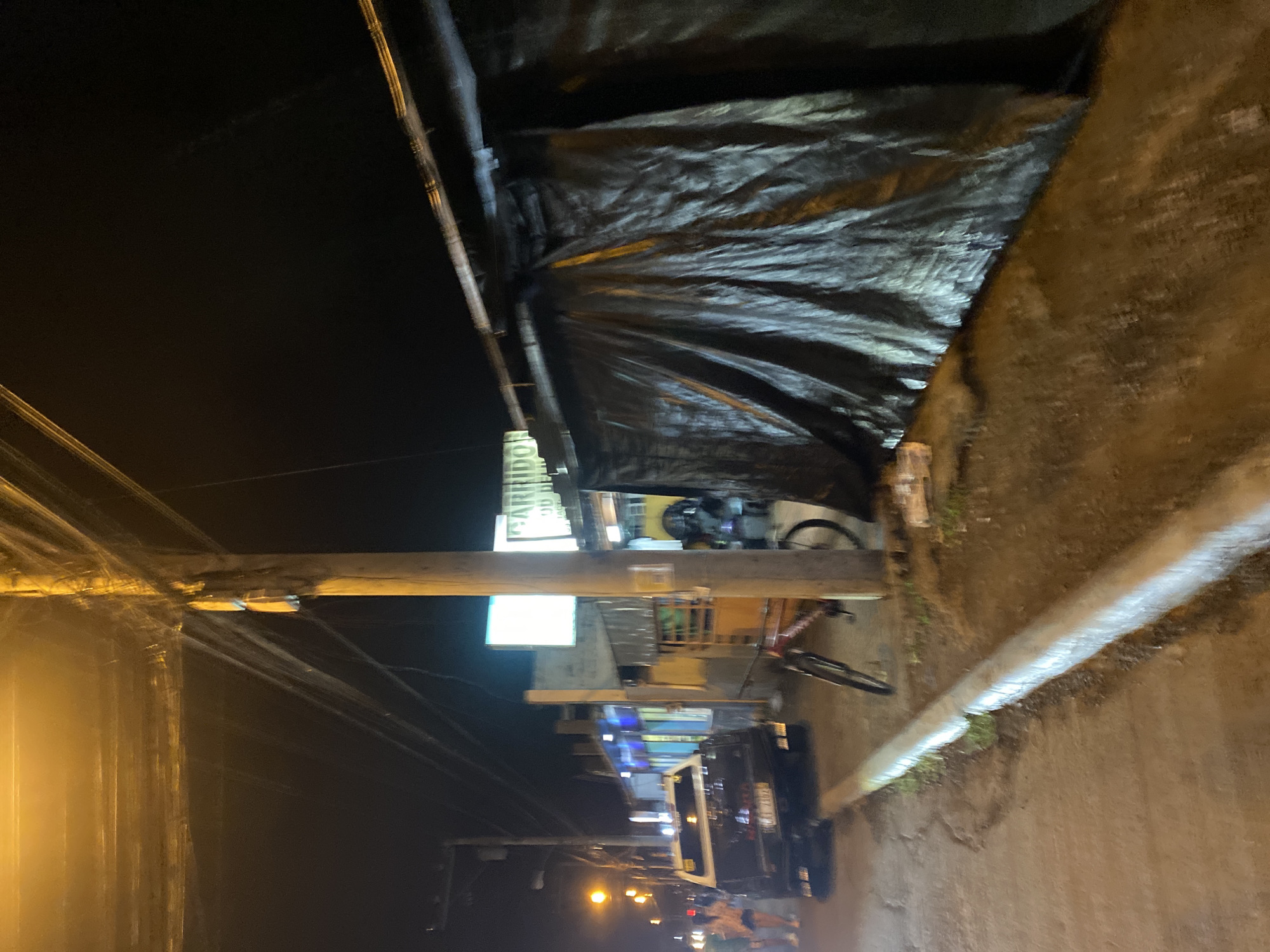 This nighttime image depicts a street scene along the Davao-Cotabato Road (National Highway) in Toril, Davao City, Philippines. The vantage point is from the ground, looking generally west down the illuminated thoroughfare.
In the foreground, a low, light-colored concrete curb or barrier extends horizontally across the lower right, separating a paved surface on the left from a darker, possibly unpaved area on the right.
To the left of the curb, a black pickup truck, identifiable by a partially visible Philippine license plate "CAT 2152," is parked facing away from the viewer. Two individuals are observed walking past the truck on the left side of the road. The area is lit by several streetlights casting a warm, yellow-orange glow.
In the mid-ground on the right, a prominent wooden utility pole rises, densely covered with numerous overhead wires. Adjacent to the pole, a dark, possibly black or navy blue, tarp or canvas sheet is stretched across a structure, creating a covered facade. A red-framed bicycle is visible, positioned near the utility pole.
The background features several commercial buildings. A brightly lit sign, prominently displaying "GARRIDON" in green lettering against a white background, with "JUAN DE LA CRUZ" below it, identifies "Garridon Food House," which is a key landmark confirming the location in Toril, Davao City. To the left of this sign, another bright white rectangular sign is visible. Further down the street, additional establishments are discernible, characterized by their blue and white artificial lighting. The sky above is dark, indicating the absence of daylight.