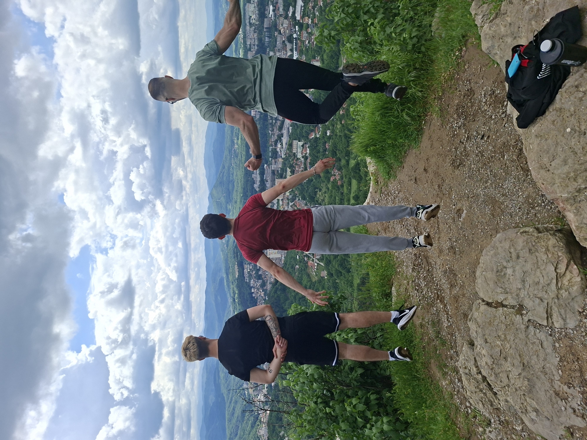 Three men are standing on a rocky outcrop overlooking Gabrovo, Bulgaria, with a cloudy sky above. The man at the top is wearing a green t-shirt and black pants and is in mid-air with arms outstretched and one leg bent. The second man is wearing a red shirt and gray pants, standing with arms outstretched. The third man is wearing a black shirt and black shorts with white stripes and is standing with his hands behind his back. The landscape below the outcrop includes green trees, mountains, and parts of the city of Gabrovo. A black bag and a water bottle are near the edge of the outcrop.