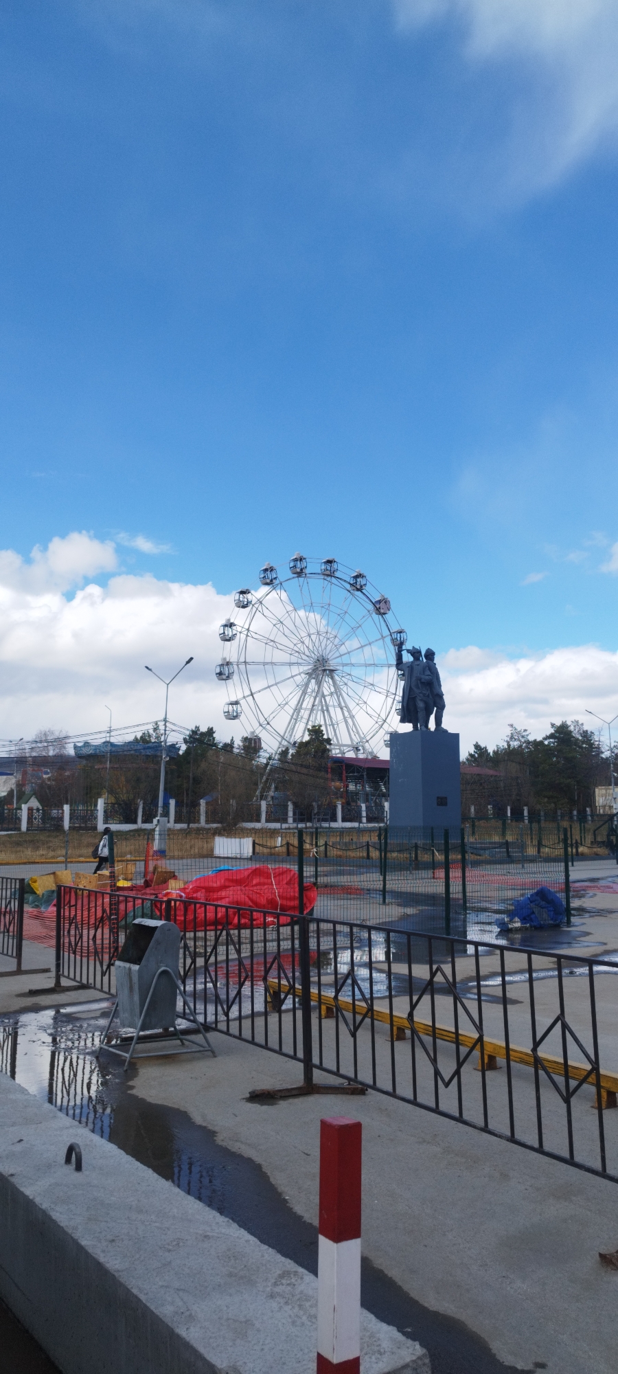 The image shows a scene in Yakutsk, Russia. A large Ferris wheel stands behind a monument depicting three figures. Both are enclosed within a fenced area. A concrete structure borders the bottom left. Puddles of water are visible on the ground. Construction materials covered in red tarpaulin and blue tarpaulin are also present within the fenced area. The sky is blue with scattered clouds. A street lamp is visible in the background.