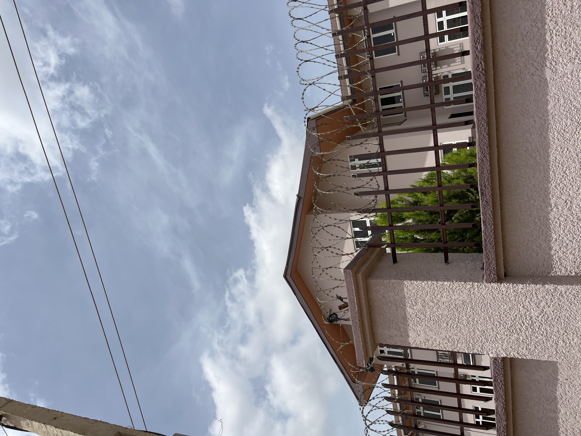 The image depicts a building in Bukuru, Nigeria, viewed from behind a fence topped with barbed wire. The building, painted a light pink or beige, has multiple windows with white frames. A section of a dark brown roof with visible wooden beams is visible at the top of the building. In front of the building and behind the fence, a dense green evergreen tree is partially visible. The fence is made of dark brown metal bars and a rough, textured pinkish-beige wall. A concrete utility pole with wires is visible on the left side of the image. The sky is partly cloudy with a bright, diffused light, suggesting daytime. There are no people visible in the image.