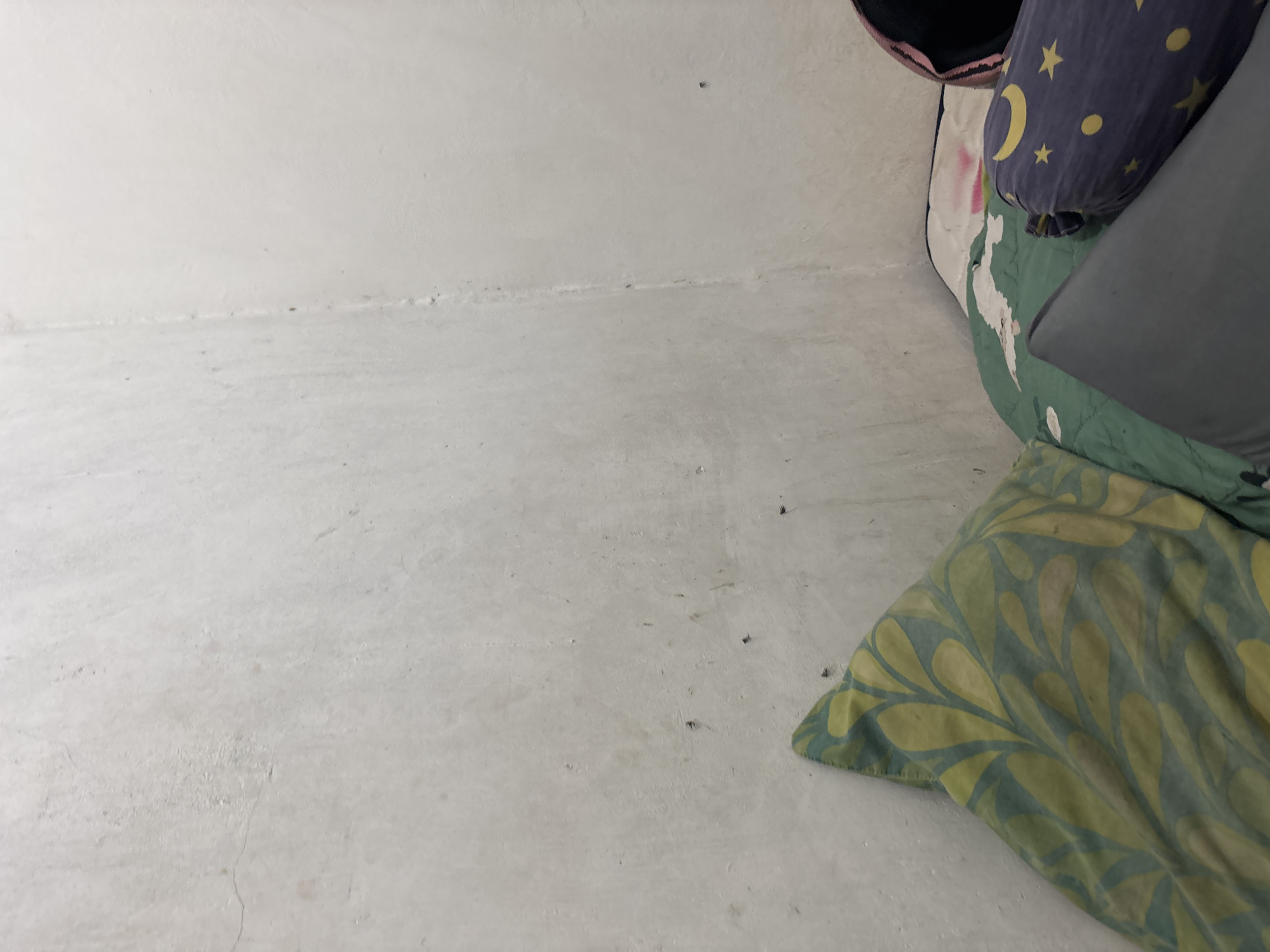 A low-angle, full shot shows a corner of a room with a bed and a textured white wall. The bed is covered with green and white patterned bedding. A dark blue bolster pillow with yellow crescent moons and stars is on the bed, to the right of the green and white patterned pillows. A dark bag is visible behind the bolster pillow. The white wall has faint marks and some cracks. This scene appears to be in a bedroom in North Jakarta, Indonesia.