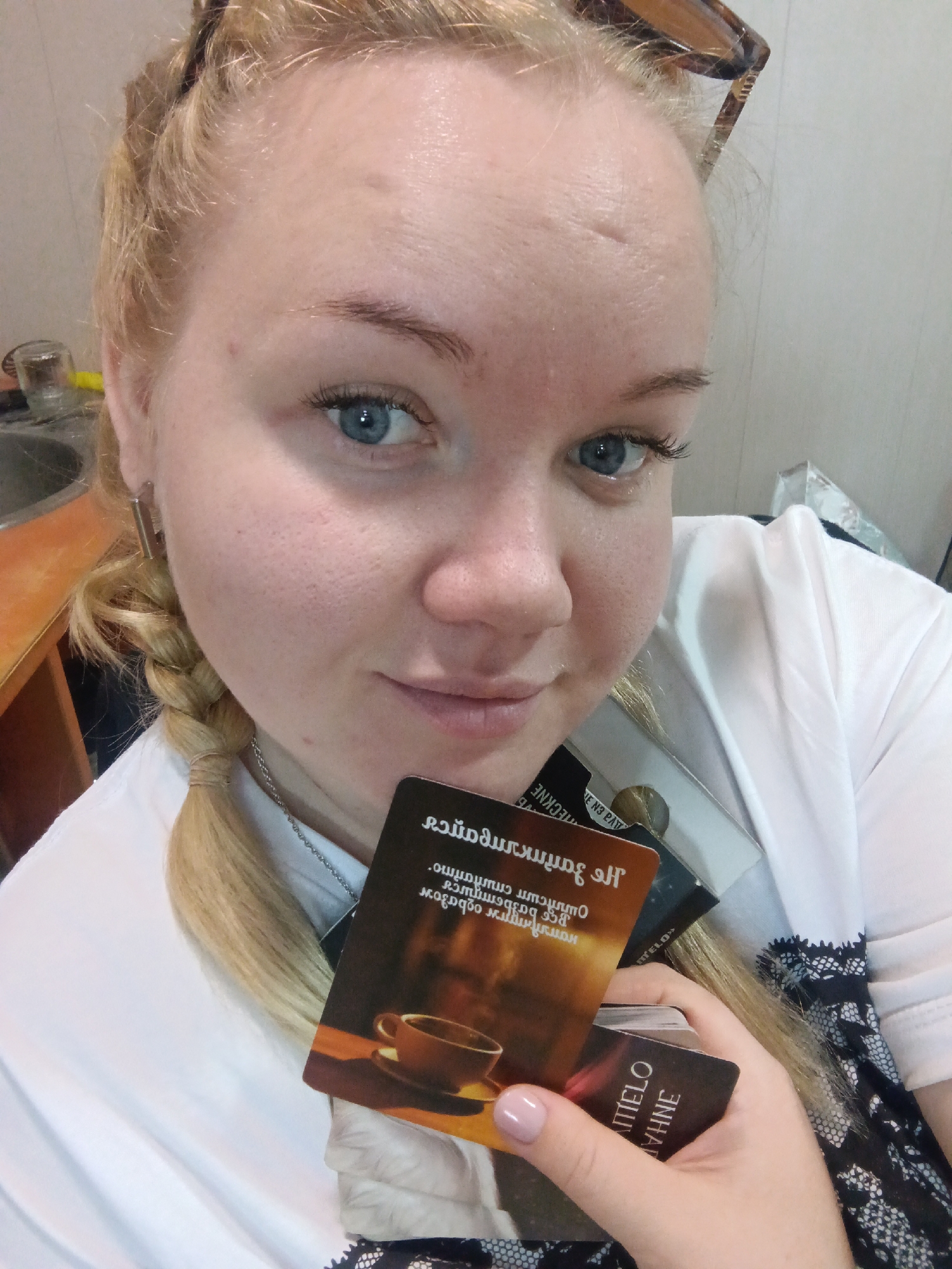 A selfie taken indoors shows a fair-skinned woman with blonde hair braided on the left side of her face. Her blue eyes are visible, and she has glasses perched on her head. She wears a white t-shirt and a silver necklace. She holds a stack of cards in her hand, one of which features an image of a cup of coffee. The text on the cards is written in reverse. The background shows a cluttered interior with a sink and a cabinet visible. There is no information available to determine if the location is Фоки, Russia.