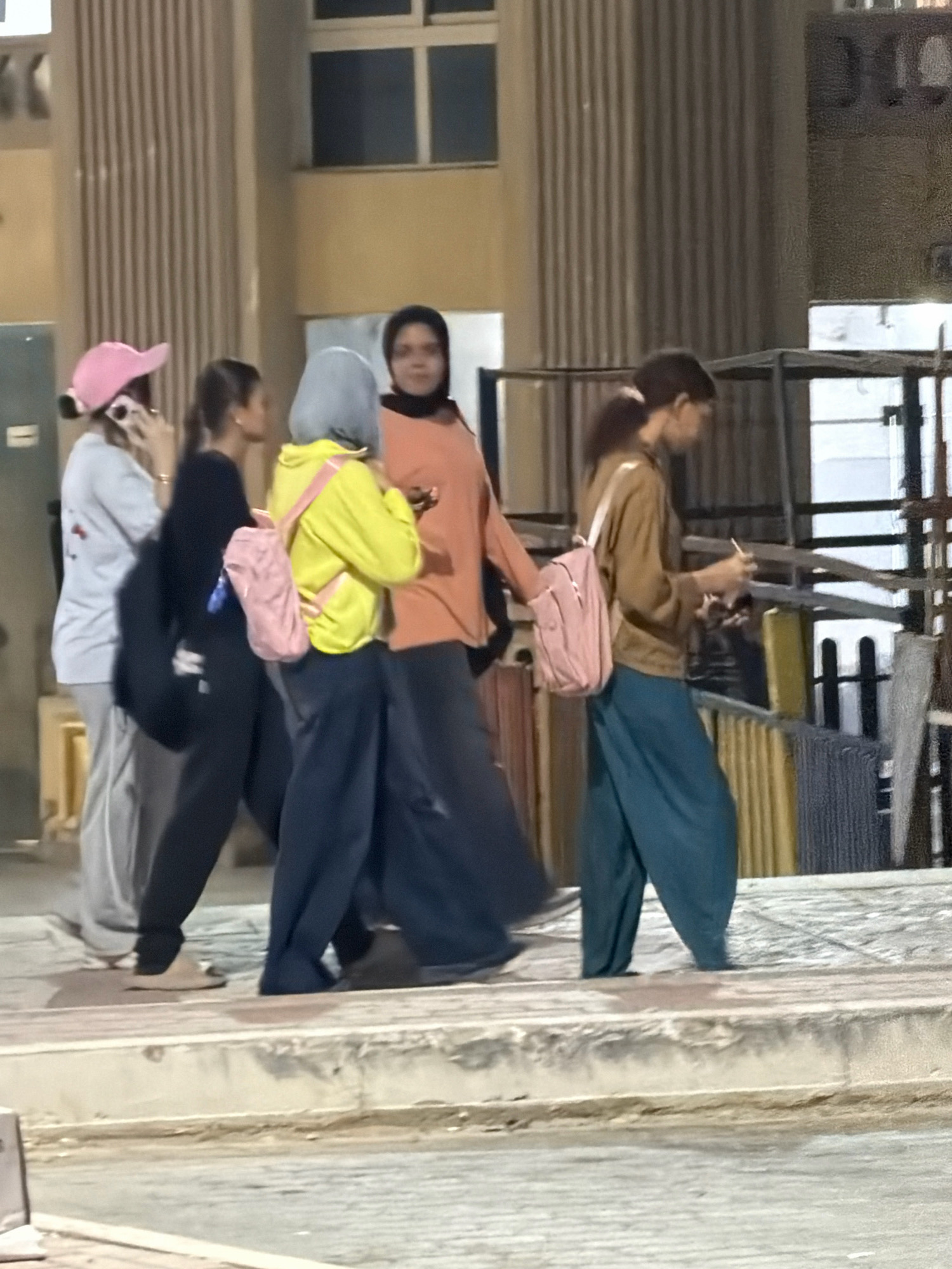 In Sharm El Sheikh, Egypt, a group of five young women are walking along an outdoor sidewalk. From left to right, the first woman is wearing a white long-sleeved shirt, grey pants, and a pink baseball cap. She is holding a phone to her ear. Next to her, a woman in a black long-sleeved shirt and black pants walks with a dark backpack slung over one shoulder and a pink backpack at her hip. The third woman, in a yellow hooded sweatshirt and dark pants, carries a pink backpack. She is looking to her left. The fourth woman, wearing an orange long-sleeved top and dark pants, walks in the same direction as the others. The last woman on the right is dressed in a brown long-sleeved shirt and teal, baggy pants. She has a pink backpack and is looking down at something in her hands. They are walking past a building with vertical brown panels and large windows. Behind them, a metal structure with shelves holds various items, some of which appear to be textiles. The ground is paved with concrete and stone.