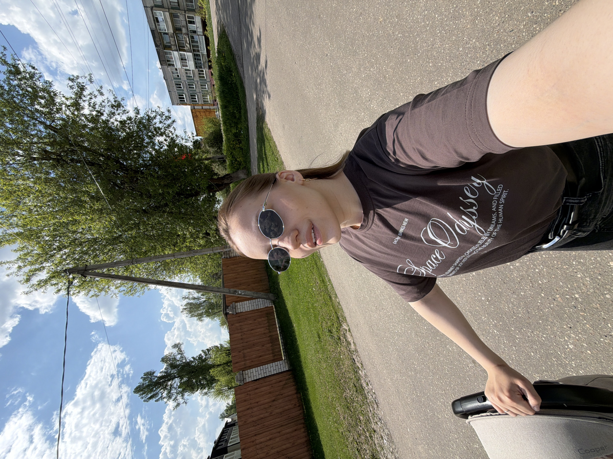 A medium-shot, low-angle, rotated selfie shows a person wearing a brown t-shirt and sunglasses, walking on a paved surface in what appears to be the city of Reshetnikovo, Russia.
The individual is fair-skinned and wears round sunglasses. The brown t-shirt features a design with the word "Space" followed by "Odyssey" in script. Text accompanying the design reads, "EACHY BRINGS NEW" and "JOURNEY SHAPED CHOICES FUELED BY DREAMS, AND FILLED OF JOY, REFUSING THE BEAUTY OF THE HUMAN SPIRIT."
The person is pushing a stroller. The brand name "Cooper" is visible on the stroller's lower right corner.
The background features a sidewalk, grass, a low wooden fence, trees, a blue sky with scattered clouds, and multi-story residential buildings. Power lines stretch across the sky. The photograph appears to be taken in daylight.