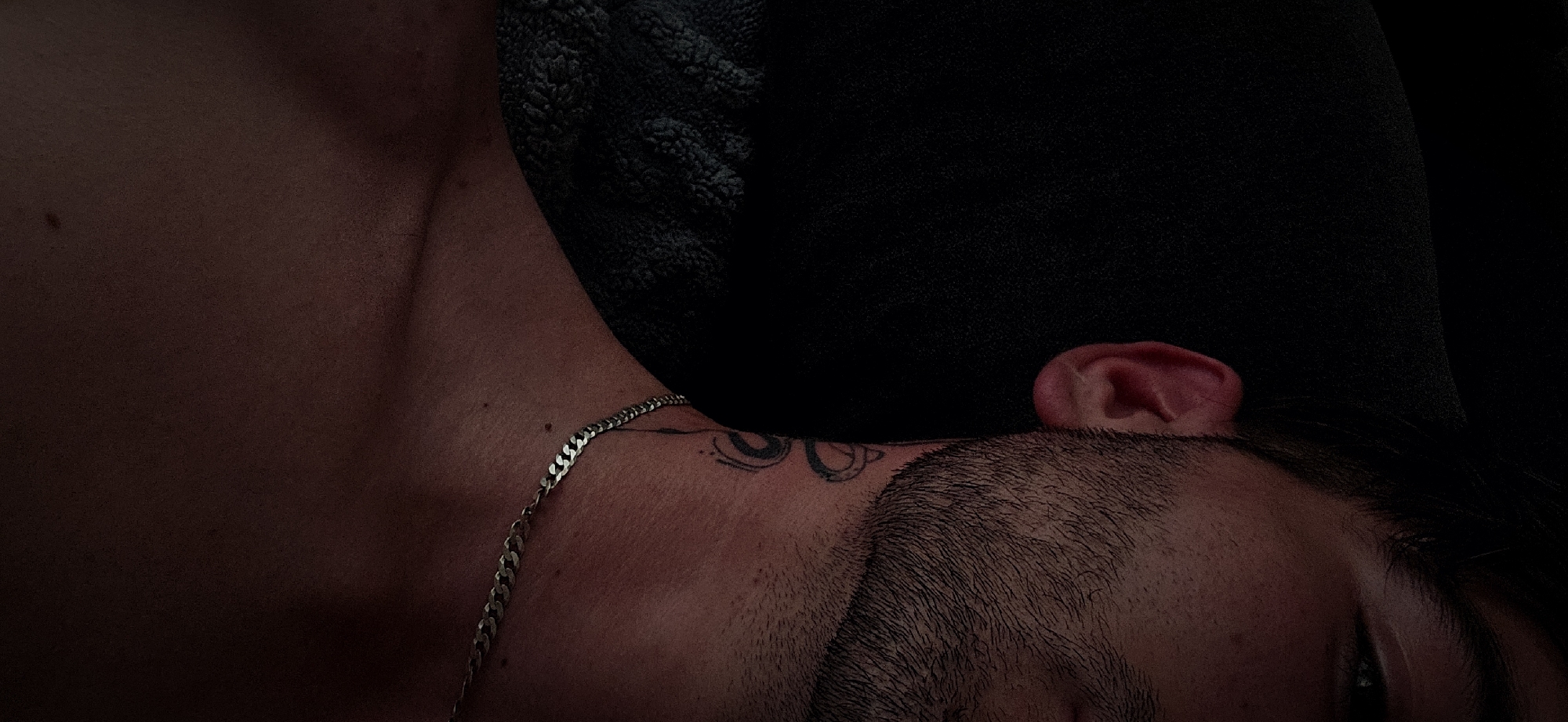 A low-angle, close-up shot depicts a person lying on their side, partially obscured by a dark, textured object, possibly a pillow or blanket. The individual's face is mostly out of view, with only the top of their head and an ear visible. Dark hair is present, along with some facial stubble. A thin, silver chain rests on their upper chest and neck, and a tattoo is visible on the side of their neck. The scene is dimly lit with predominantly muted, reddish-brown tones. The image contains no specific indicators to place the location within Plovdiv, Bulgaria.