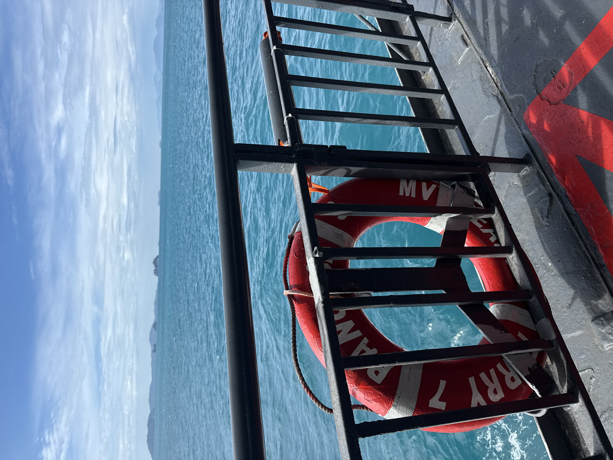 A red and white life preserver hangs on a metal railing overlooking the turquoise ocean on a sunny day. In the distance, islands with green tops are visible against a pale blue sky dotted with white clouds. The railing is black and appears to be part of a ferry or boat. The deck below is gray with a red arrow painted on it. The water is slightly choppy. The scene appears to be in Thailand, possibly near an island destination.