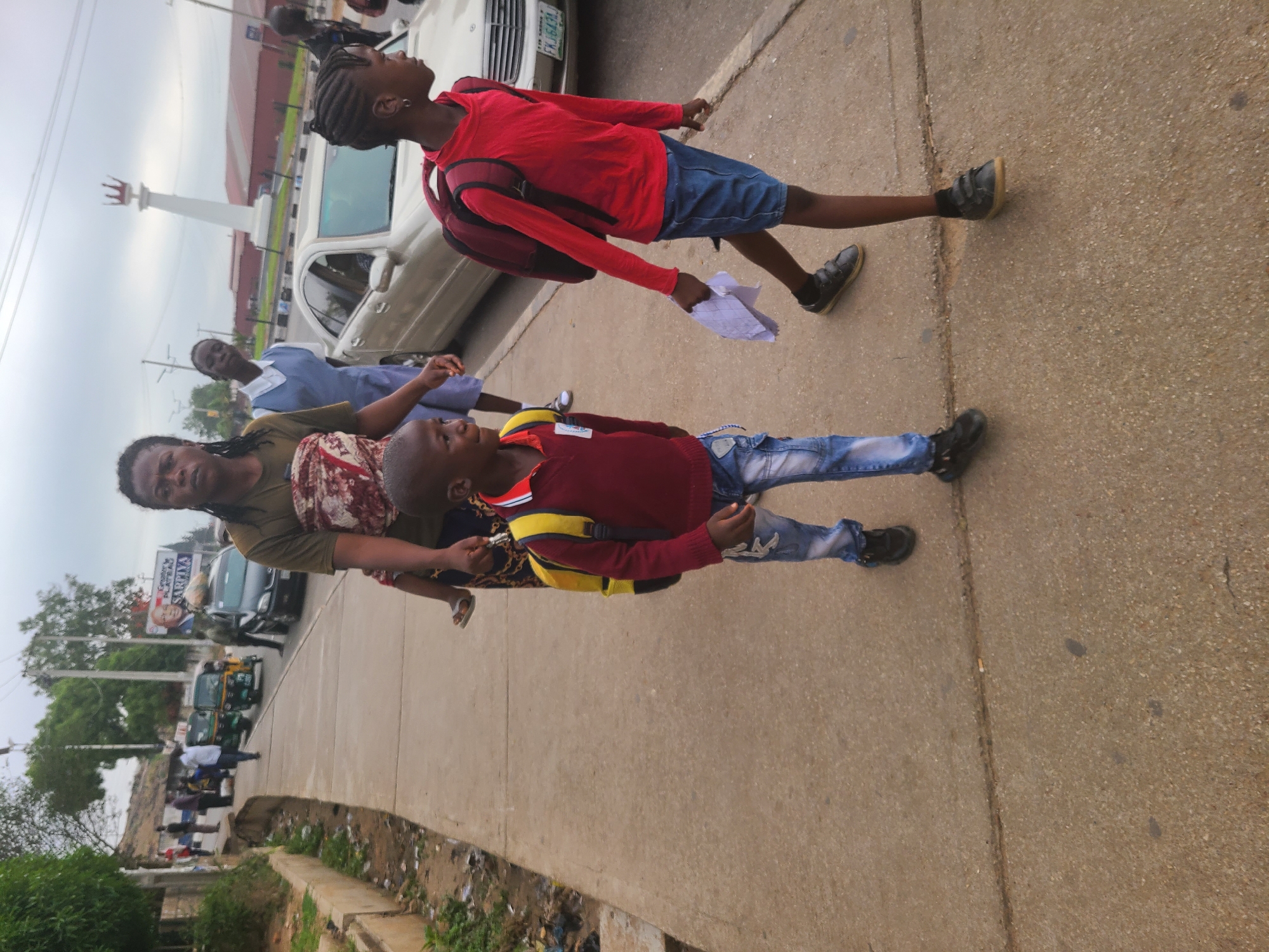 The image shows a group of people walking on a sidewalk in Jos, Nigeria. The foreground features a woman with two children, one older and one younger. The older child wears a red shirt and blue shorts with a maroon backpack, while the younger child wears a similar red shirt with ripped blue jeans and a yellow backpack. All three are walking away from the camera.
Behind them, there is a woman in a blue uniform. A white car is parked to the side with a partial view of the license plate. In the background, there is a white monument and other vehicles. A sidewalk edge is lined with some vegetation and trash. There appear to be pedestrians along the edge of the roadway as well.