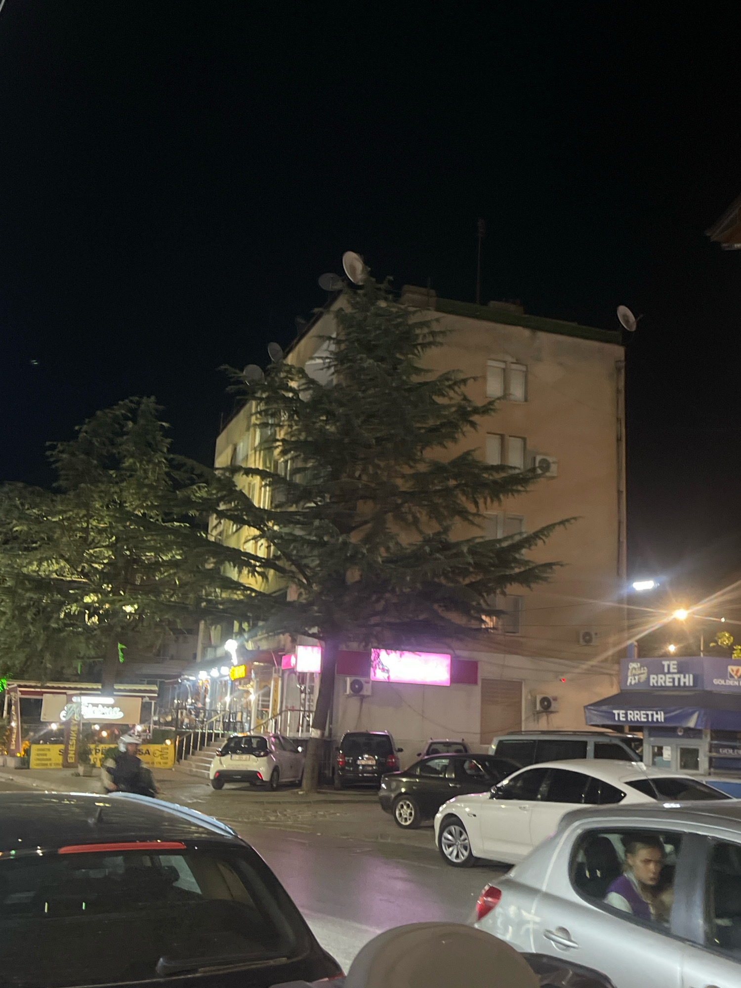 An exterior nighttime shot shows a street scene in Prizren, Kosovo. A multi-story building with several satellite dishes on the roof and an illuminated facade is visible in the background. Trees in front of the building partially obscure it. Several cars are parked and driving on the street, including a black sedan in the foreground, a white sedan, a small white car, and a darker sedan. A person wearing a helmet is riding a motorcycle. Buildings with illuminated signs line the street. A person is visible inside the silver car in the foreground. The overall lighting suggests an urban environment at night, with streetlights and building lights illuminating the scene.