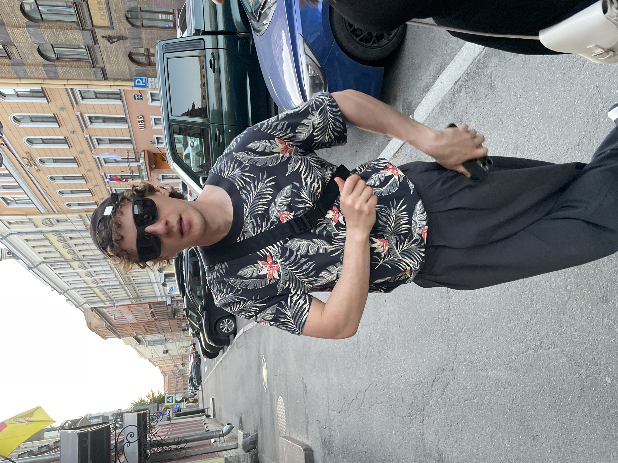 This image captures an urban street scene in Saint Petersburg, Russia, during daytime under bright conditions.
In the foreground, a young to middle-aged male with brown, curly hair and light skin tone stands facing the camera. He wears black rectangular sunglasses, a black short-sleeved t-shirt with a white, grey, and red tropical floral pattern, and dark, likely black, pleated pants. A black crossbody bag with a wide strap is slung across his chest. His left hand rests on the bag's strap, while his right hand holds a dark-colored smartphone, partially inserted into his right pants pocket. His mouth is slightly open, and he maintains a neutral expression.
To the far right, partially obscured by the main subject, is another individual whose dark clothing and a light-colored (possibly cream or white) crossbody bag are visible.
The scene is set on an asphalt street with visible cracks and two circular manhole covers. A sidewalk runs along the left side of the street, and numerous parked cars line the right side. These include several black SUVs, a dark green vehicle resembling a UAZ Patriot, and a blue car, possibly a Hyundai Solaris or Accent.
Both sides of the street are flanked by multi-story historical buildings of varying pastel hues, including yellow, orange, and brown brick. The building on the far left features a distinct cornice and displays a yellow flag with horizontal stripes and a coat of arms, resembling the flag of Moldova. Further down on the right, an orange building with arched windows is visible, featuring a small balcony from which a Russian flag (white, blue, red horizontal stripes) and a white flag hang. This building also prominently displays a sign that reads "ОТЕЛЬ" (Hotel) in Cyrillic. Adjacent to it, a brick building with arched windows features a partial sign reading "ОБЩЕСТВА РЫБО..." (likely "Society of Fishermen" or similar). Street signage includes a blue parking sign with the letter "P" and "8:00 - 20:00" in Cyrillic, and a yellow triangular pedestrian crossing sign. Drainpipes are visible on the facades of the buildings.
The sky overhead is bright and clear, indicating a daytime setting, possibly late afternoon or early evening, with sunlight causing some glare on the upper left portion of the image.