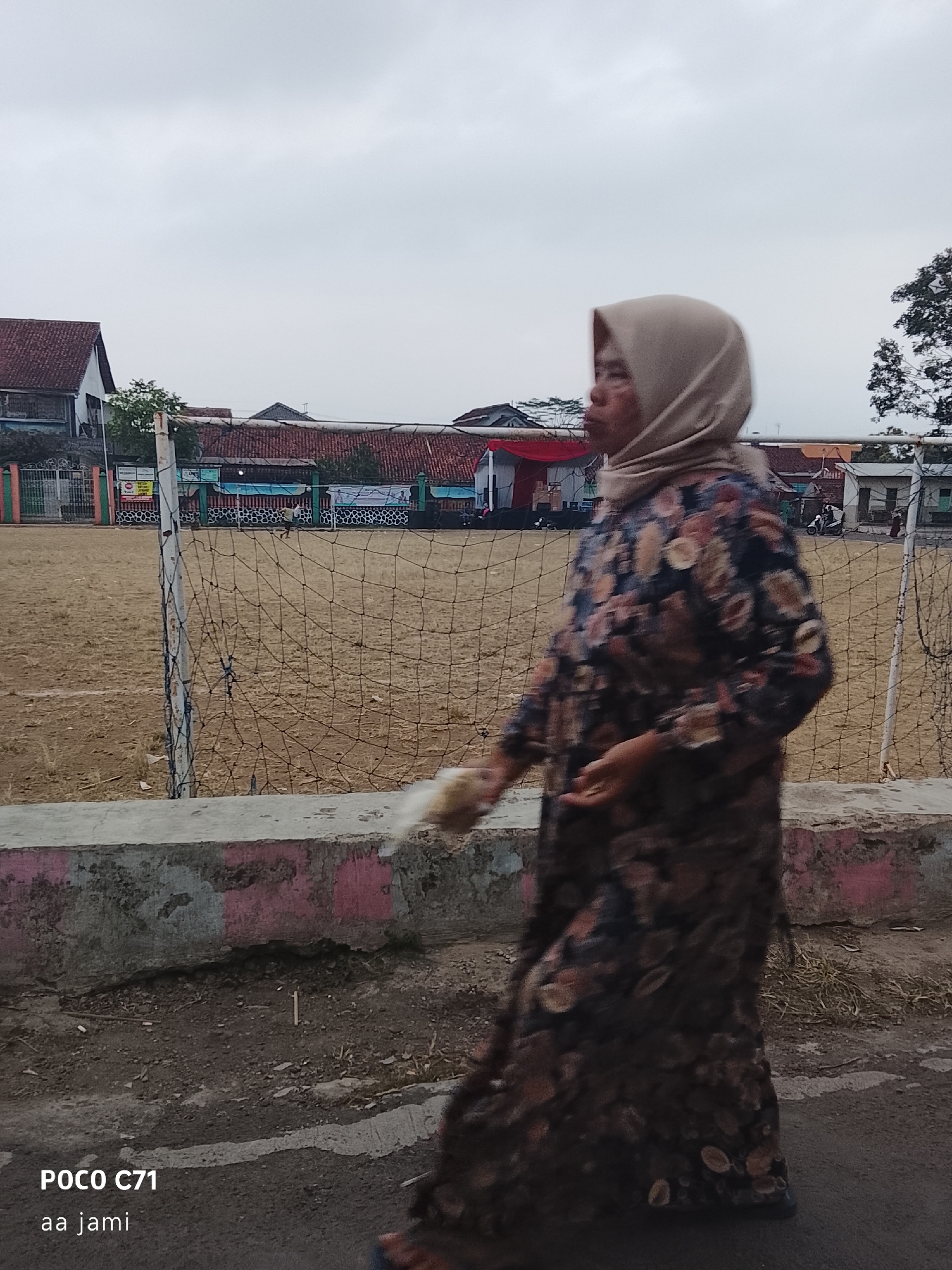 A full-body, side-profile shot captures a woman walking in front of a dusty field with a volleyball net. The woman, seen from the chest down, is wearing a light brown hijab and a long, dark floral-patterned dress. Her left hand is visible, holding a small white object. The background depicts a fence surrounding the field, with buildings and trees visible beyond it. The sky is overcast. The image includes text in the lower left corner indicating "POCO C71" and "aa jami". The location is implied to be in Tasikmalaya, Indonesia, based on the user's prompt, though there are no explicit visual cues to confirm this.