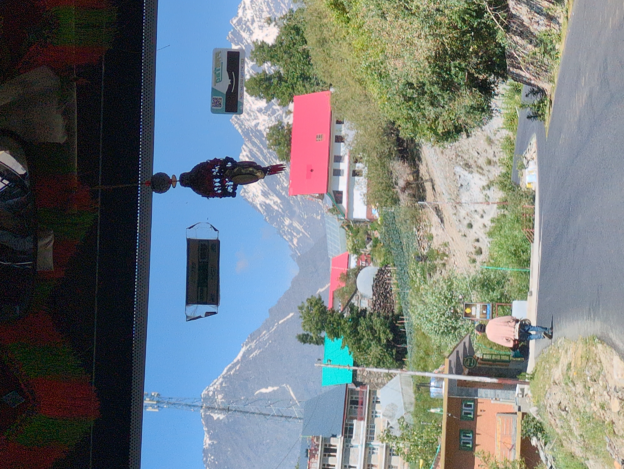 The image captures a street scene in Khangsar, India, with a prominent mountain range in the background. The mountains are snow-capped, indicating a high altitude. In the foreground, a paved road winds through a village. To the left of the road, a man wearing a light-colored jacket and jeans walks away from the camera, carrying an object. Beside the road, there are several buildings with various colored roofs, including red and teal. Trees and vegetation are scattered throughout the scene, with some areas appearing terraced, possibly for agriculture. A telecommunications tower is visible on the left side of the frame, rising above the buildings. Two rectangular objects are suspended from the top of the frame, appearing to be some form of signage or decoration. The sky is clear and blue.