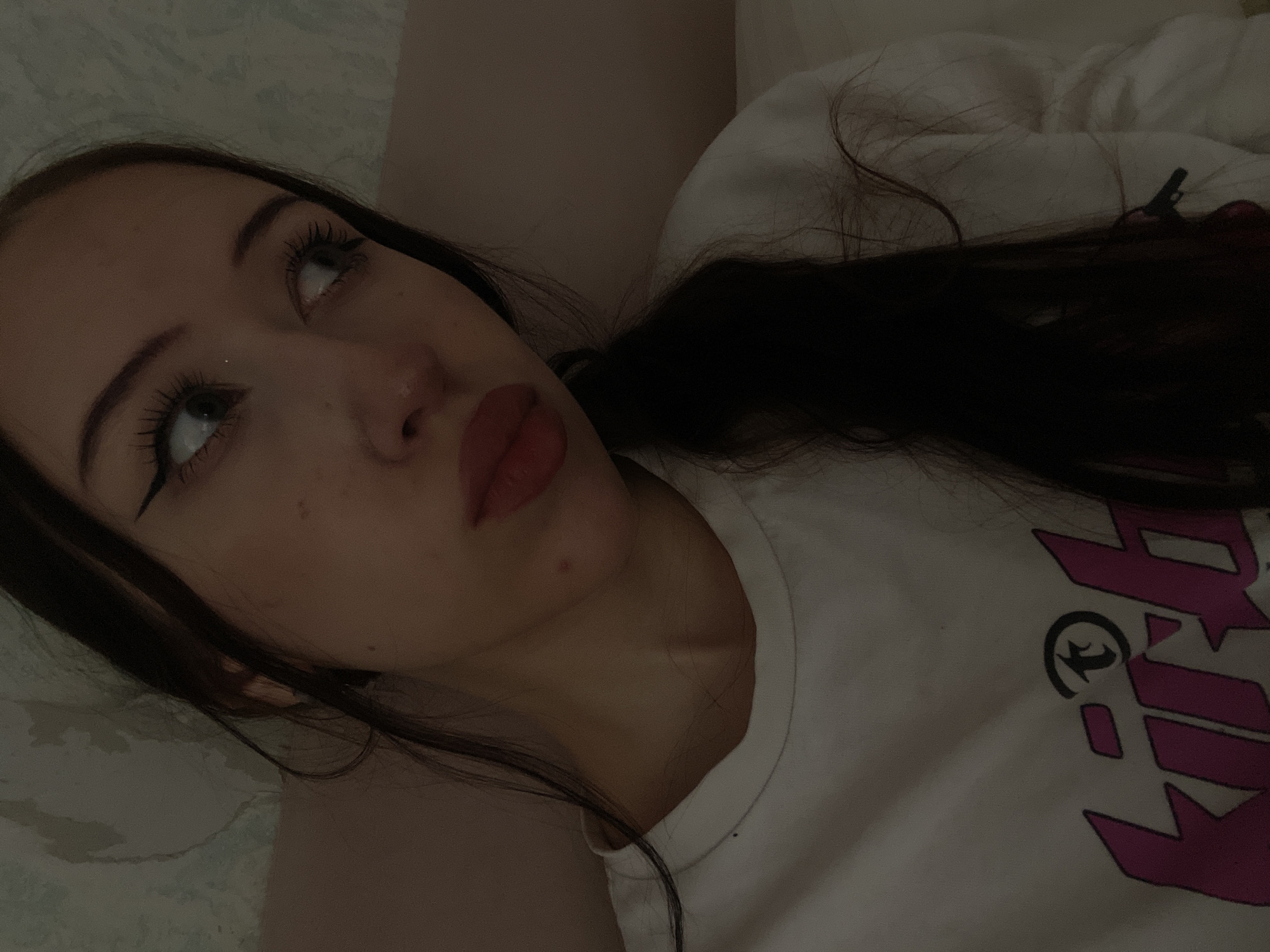 The media file is a close-up, overhead shot of a young woman lying down. Her eyes are open and looking upwards, with heavy eyeliner and mascara applied. Her lips are painted a deep red. She has dark hair that is spread out around her. She is wearing a white t-shirt with a bold, pink graphic design on it. The graphic appears to be lettering or a logo. The setting is dimly lit, possibly indoors, with a soft, textured surface beneath her, suggesting a bed or couch. The mood conveyed is contemplative or introspective. There are no visible environmental cues to suggest a specific time of day or weather. The location context provided is Votkinsk, Russia.
