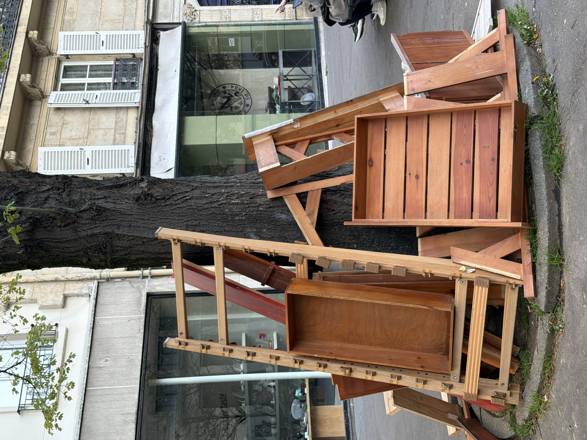 The image shows discarded wooden furniture and frames piled on a sidewalk in Paris, France. The objects lean against a large tree. The backdrop includes buildings with shuttered windows and storefronts with glass facades, one of which displays a logo with "ON TAP EST 1884". A person's legs and feet are visible near the top of the frame. The sidewalk is concrete with grass growing along the edge.