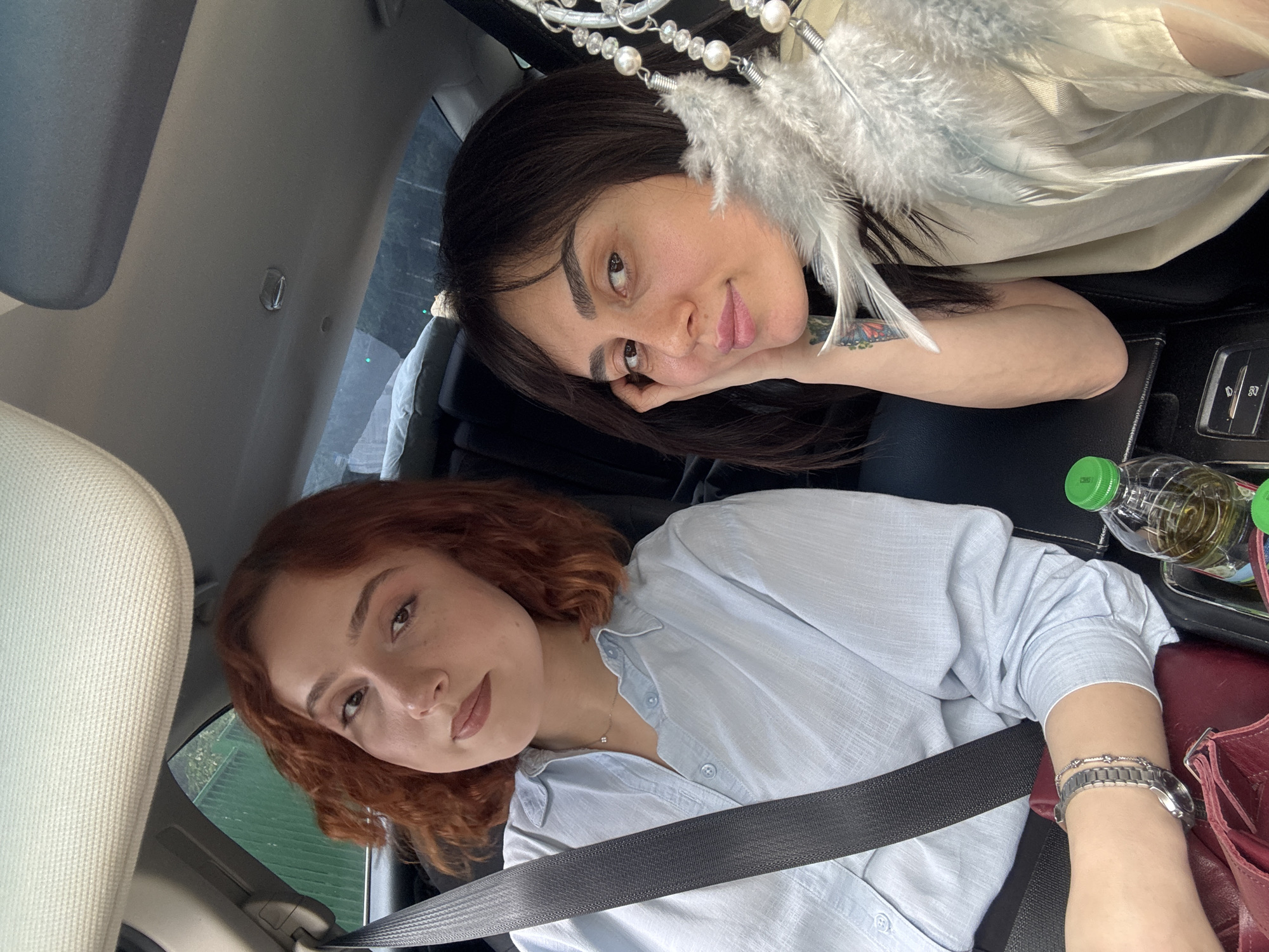 Two women are posing in a car. The woman on the bottom has red curly hair, is wearing a light blue button-down shirt, and has a seatbelt on. She also wears a watch on her left wrist. The woman on the top has dark hair and rests her head on her hand. A dream catcher hangs near her. The car interior is visible, including a water bottle, part of a red bag, and interior features. The location is Tbilisi, Georgia, evidenced by the type of vehicle and architectural elements of the background seen through the car window.