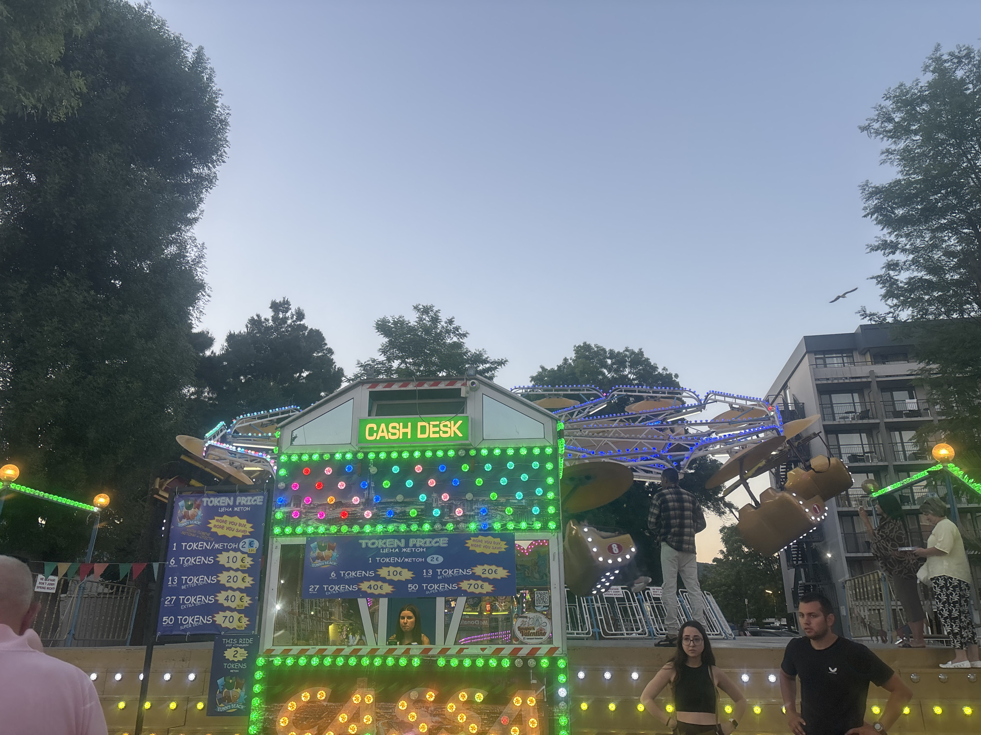 In Nesebar, Bulgaria, a scene unfolds at a carnival or amusement park. A brightly lit "Cash Desk" sign illuminates a booth adorned with colorful lights. A sign displays "Token Price" with various token package options and their corresponding prices in Euros. Inside the booth, a young woman appears to be serving customers. Outside the booth, several people are visible. In the foreground, a man and a woman stand with their hands on their hips, looking towards the camera. To the right of the booth, another man is standing with his hands behind his back, looking forward. Further back, a woman in a yellow top and patterned skirt interacts with another individual. In the background, an amusement park ride with yellow seats is in motion, and people are visible on and around it. Trees frame the scene, and a multi-story building with windows is visible in the upper right. The sky is a pale blue, indicating either early morning or late evening.