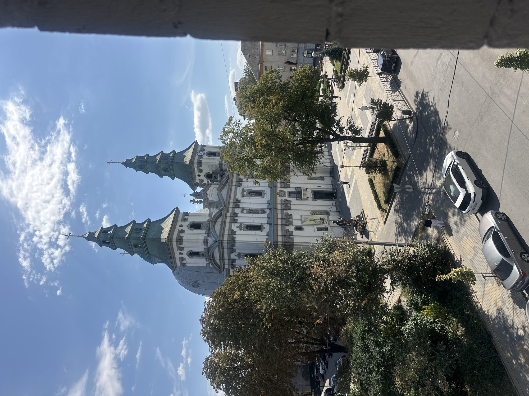 This image captures a view of the Ternopil Theological Seminary and the Church of the Transfiguration of Jesus in Ternopil, Ukraine, from a higher vantage point, likely a window. The dominant features are the twin green spires of the church, topped with crosses, and the white facade of the seminary building with its classical architectural details.
In the foreground, the edge of a stone structure, possibly a window frame or balcony, partially obscures the view on the right. Below the seminary and church, a street is visible with several cars parked and driving. Among them, a silver sedan is parked on the left side of the street, and a white modern SUV is driving away on the right. People are seen walking on the sidewalks and in the plaza area in front of the church. Trees with green and some autumnal foliage are scattered throughout the scene, casting shadows on the pavement. The sky is blue with scattered white clouds, indicating a bright, sunny day. The overall scene depicts a typical urban setting with a prominent religious and educational institution.