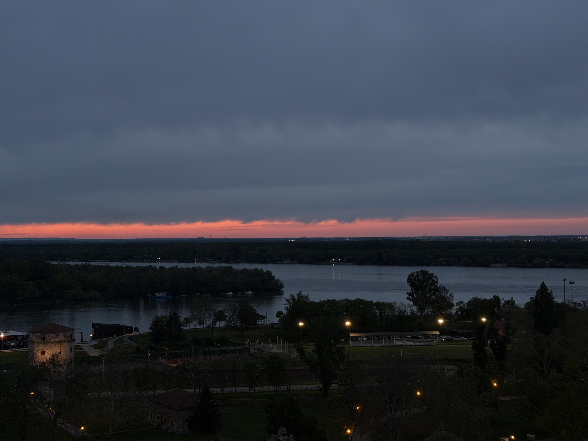 This image captures an elevated view of a river landscape at dusk in Belgrade, Serbia. The scene is dominated by a dramatic sky featuring a broad band of dark, heavy clouds covering the upper two-thirds of the frame. Below these dark clouds, a striking orange and red glow is visible along the horizon, hinting at the last light of a sunset or the first light of a sunrise, though the activated artificial lights in the foreground strongly suggest twilight or early night.
In the midground, a wide river, likely the Sava or Danube, flows from left to right, its surface reflecting the subdued light. The far bank is covered by a dense, dark forest, with hints of distant urban lights breaking through the trees on the horizon. A large, dark boat or barge is discernible on the river's surface towards the right.
The foreground depicts a park area, now mostly in shadow, with various artificial lights illuminating pathways and structures. To the left, a prominent, round stone tower, reminiscent of the Nebojša Tower, stands illuminated, showing windows and architectural details. In the center and right foreground, multiple streetlights and park lights cast pools of warm, yellow light onto paths and grassy areas. Part of what appears to be a sports field, possibly a soccer pitch, is visible on the right, also partially lit. The overall setting combines historical architecture with natural elements and urban parkland, under a captivating twilight sky. No specific human activity is discernible, and there is no visible text in the image. The mood is calm and serene, characteristic of the transition from day to night.