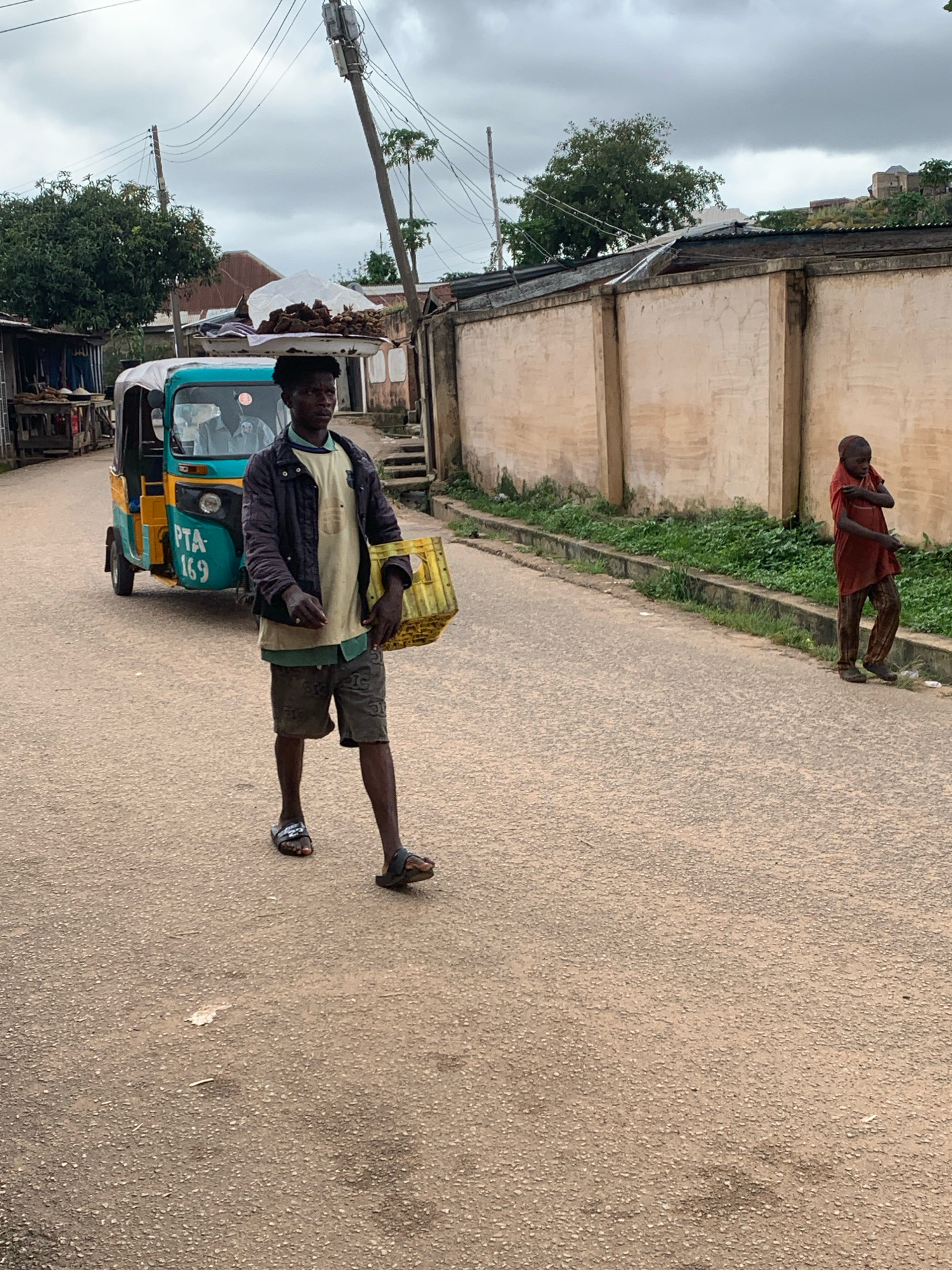 A young man wearing a dark jacket over a light-colored shirt and shorts walks down a paved street in Jos, Nigeria, carrying a yellow basket. To his left, a blue and yellow tricycle taxi with "PTA 169" written on its side is parked. Further down the street, a small child in a red shirt and patterned pants stands by a concrete wall. The sky is overcast. The area appears to be a residential or commercial street with buildings and greenery visible.