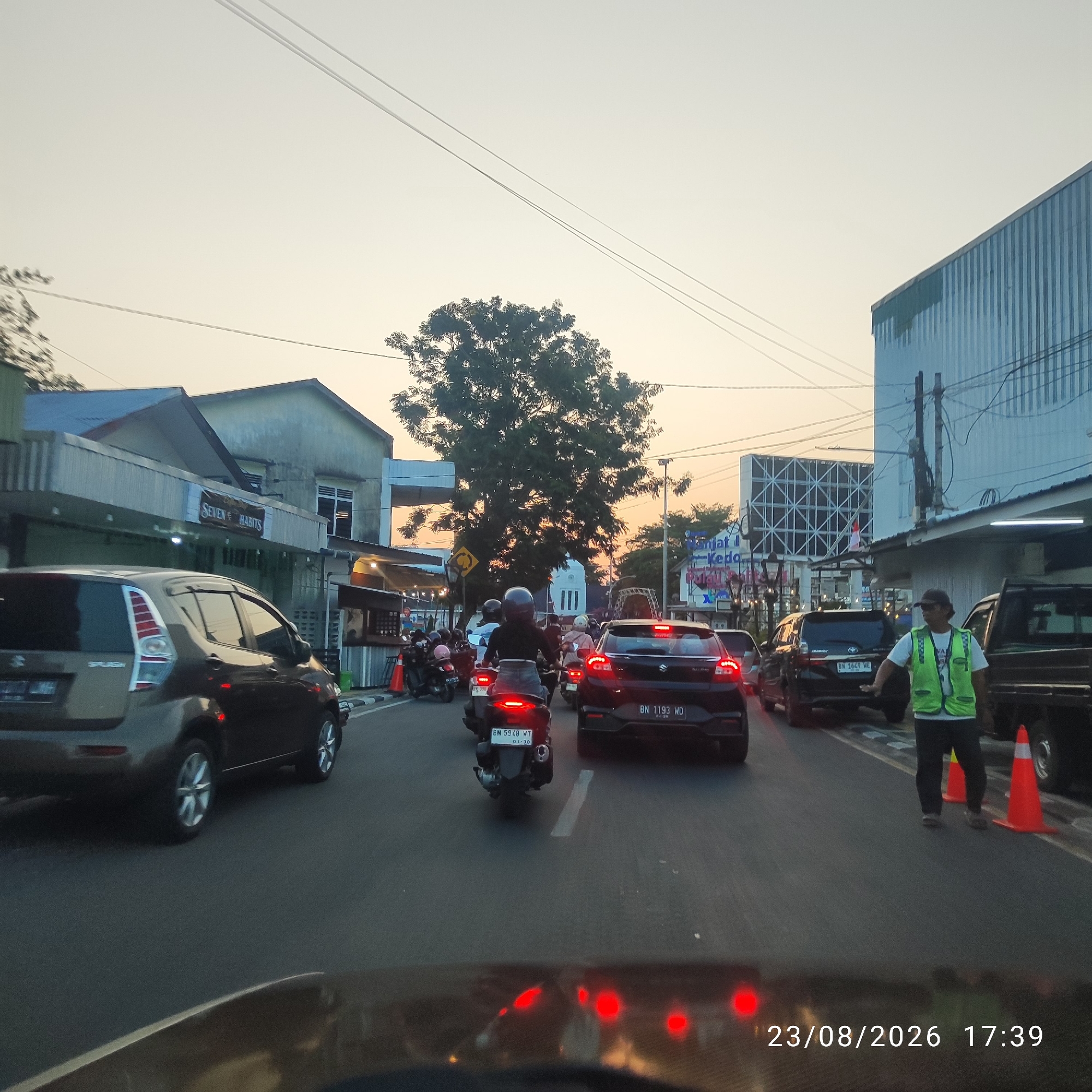 The image depicts a street scene in Tanjung Pandan, Indonesia, during what appears to be late afternoon or early evening, indicated by the warm, low light and orange hues in the sky. The view is from the perspective of a vehicle's dashboard, showing the road ahead. The street is busy with traffic, including cars and numerous motorcycles.
On the right side of the road, a traffic controller wearing a high-visibility green vest and a dark cap is directing traffic. Orange traffic cones are placed on the road near him. In the background, buildings line the street, some appearing commercial with signage, including one that reads "SEVEN HABITS." Other structures include residential buildings and what looks like a large warehouse or industrial building with a corrugated metal facade. Trees are visible, including a prominent, large tree in the middle ground. Electrical wires are strung overhead.
The timestamp "23/08/2026 17:39" is overlaid on the bottom right of the image, suggesting the date and time the photograph was taken. The overall scene conveys a sense of daily urban life and traffic congestion.