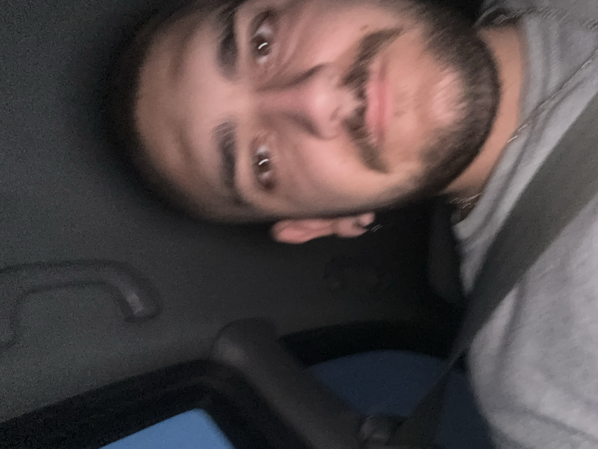 A close-up, selfie-style photograph captures a young man inside a vehicle. He has short, dark hair and a mustache and beard. He is wearing a grey t-shirt and a silver chain. The man is looking directly at the camera. The interior of the vehicle is dark, with a sunroof visible at the top left of the frame. The image appears to have been taken at night. There is no visible indication of the specific location within Staro Selo, Bulgaria.