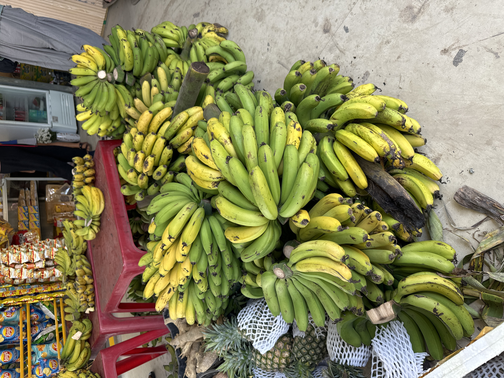 This media shows an outdoor market scene in Nam Hải, Vietnam, dominated by large quantities of bananas and other produce. The primary subjects are numerous bunches of bananas, mostly green with some starting to ripen to yellow, piled on the ground and on red plastic stools. Many bananas show natural blemishes and dark spots. Below some of the banana piles, several pineapples are visible, some of which are protected by white foam netting.
In the background on the left, a market stall is visible with various packaged goods. A white refrigerator unit can be seen, and below it, shelves stocked with bags of snacks. The "Lays" brand name is clearly discernible on several bags of potato chips. A person's lower body, dressed in dark pants, is partially visible near the snack display, suggesting the presence of a vendor or customer.
The scene takes place on a concrete surface, which appears somewhat dirty or stained, indicative of a busy outdoor environment. The lighting is bright and even, suggesting it is daytime. The activity depicted is the display of fruits and snacks for sale in a market setting. No specific human action is captured, but the arrangement implies commercial activity.
