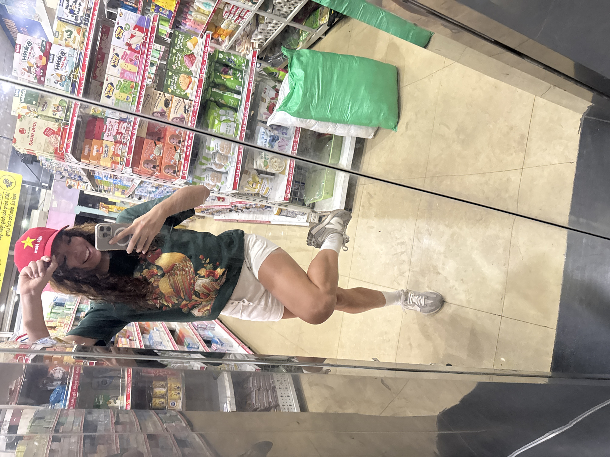A person with long curly hair, wearing a red baseball cap with a yellow star and text, a dark green shirt with a large bird graphic, and white shorts, is standing in a store and taking a mirror selfie with a silver iPhone. They are wearing gray sneakers and white socks. The store shelves are stocked with various food products and snacks. There are also large green and white sacks on the floor. A yellow sign with black text in a non-English language is visible on the shelf.