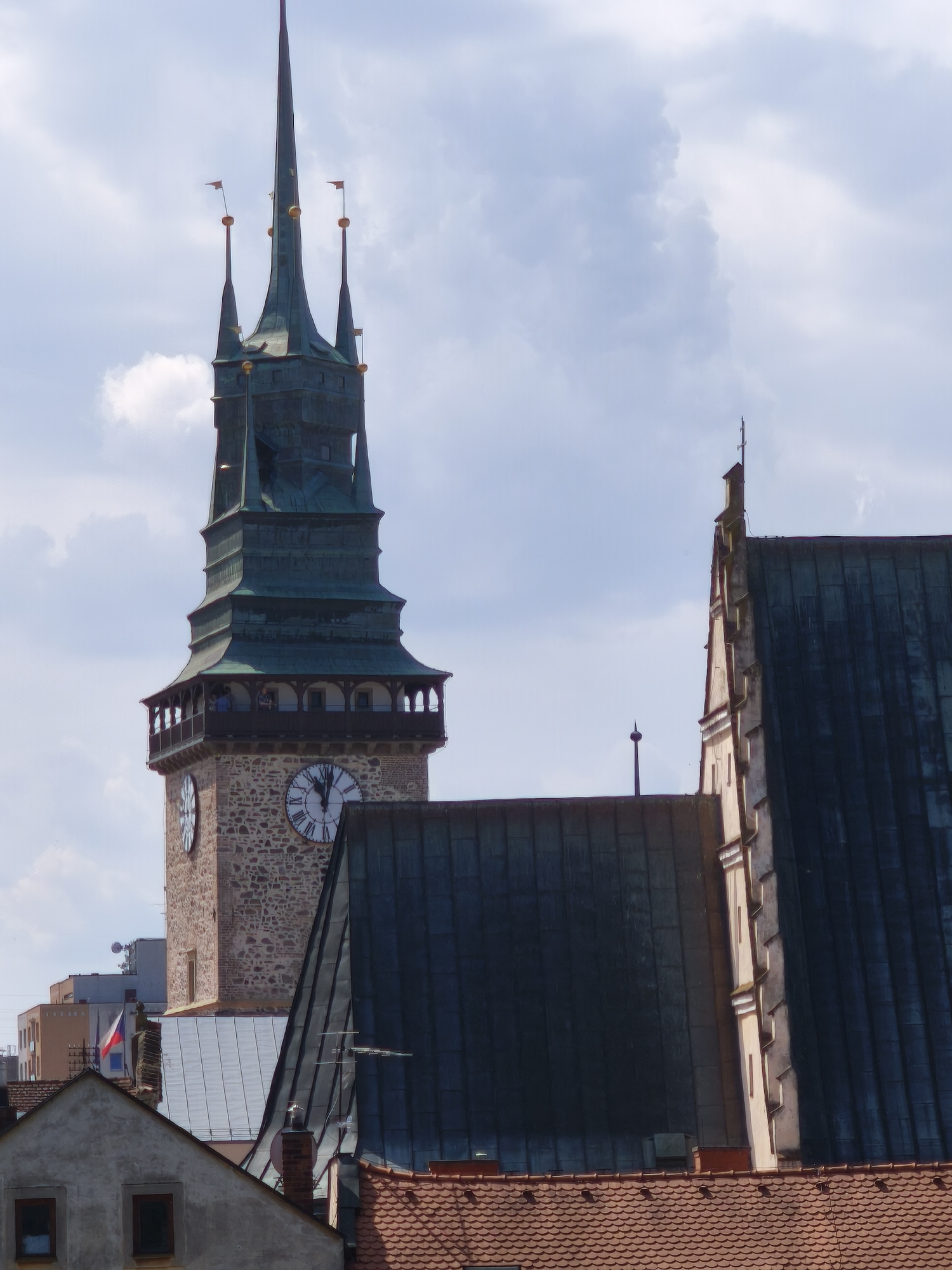 This image captures an exterior view of the Pardubice Castle in Pardubice, Czechia, focusing on its prominent clock tower and adjacent buildings. The clock tower, constructed of stone and featuring a green, multi-tiered spire topped with flags, dominates the left side of the frame. It has a balcony area with arched openings and two large clock faces visible. To the right of the tower, another building with a steep, dark, metallic roof and light-colored stone accents is partially visible. In the foreground, the tiled roofs of smaller buildings are present, with one showing a section of a house with windows. The sky above is a mix of blue with scattered clouds. Several people are visible on the balcony of the clock tower. The overall scene depicts a historic urban landscape under a cloudy sky.