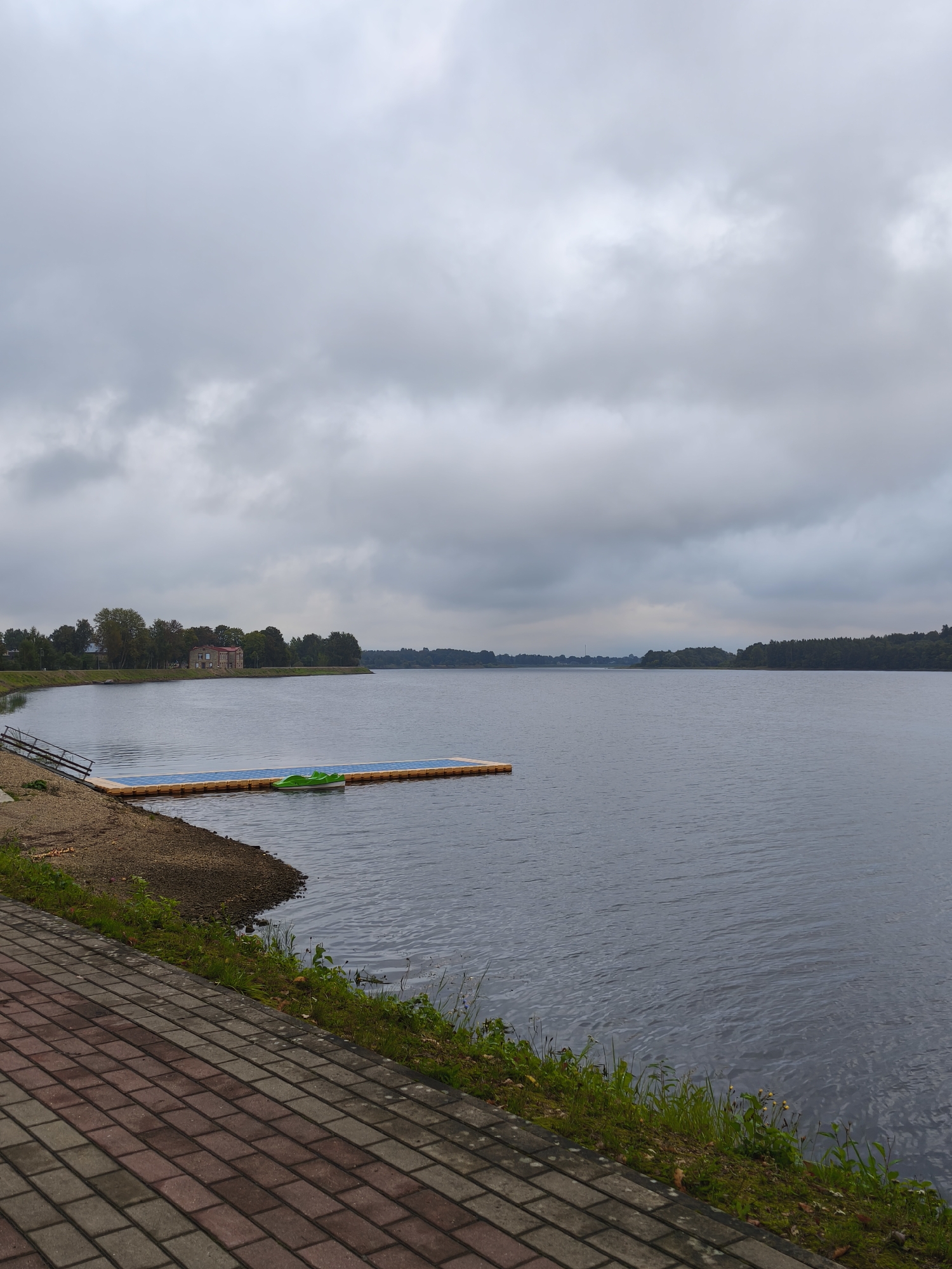 The image depicts a cloudy day at a waterfront in Pļaviņas, Latvia. In the foreground, a paved walkway made of red and gray bricks runs diagonally from the bottom left to the bottom right of the frame. Adjacent to the walkway, a strip of green grass and some small plants grows along the edge of the water.
The water itself is a dark, rippling expanse, occupying the majority of the image. On the left side of the water, a small beach area with gravel leads down to the water's edge. A wooden ramp or pier structure is visible on this beach. Further out in the water, a floating platform is anchored. This platform appears to be composed of modular, buoyant units and has blue surfaces. A bright green paddle boat is docked on the edge of this platform.
In the middle ground, across the water, is the shoreline of Pļaviņas. It is lined with a dense row of trees, beyond which a large, multi-story brick building with a dark roof is visible. The sky above is overcast with heavy, gray clouds, suggesting a cool or potentially rainy day. The overall atmosphere is somber and tranquil.