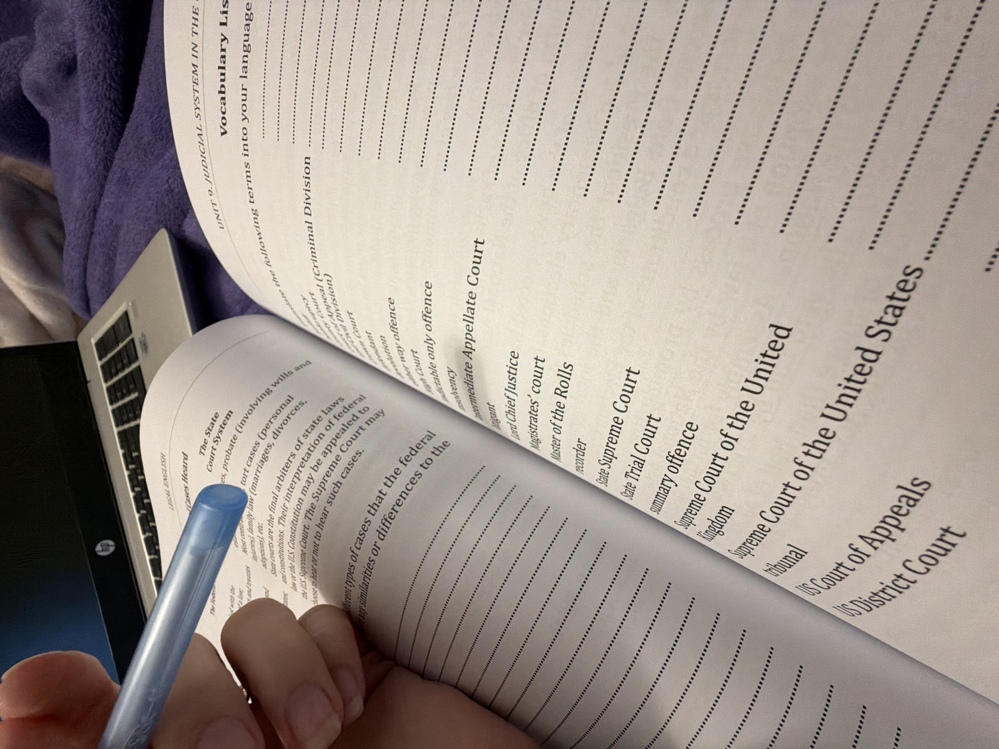 The image shows a person holding a light blue pen and studying a vocabulary list for Unit 9: Judicial System. The vocabulary list includes legal terms such as "Intermediate Appellate Court", "Supreme Court of the United States," "State Trial Court", "Master of the Rolls" and "summary offence", among others. The book appears to be open on a surface next to a partially visible laptop computer. The scene is likely indoors, possibly at home, and does not contain any specific indication of being located in the city of Plovdiv, Bulgaria.