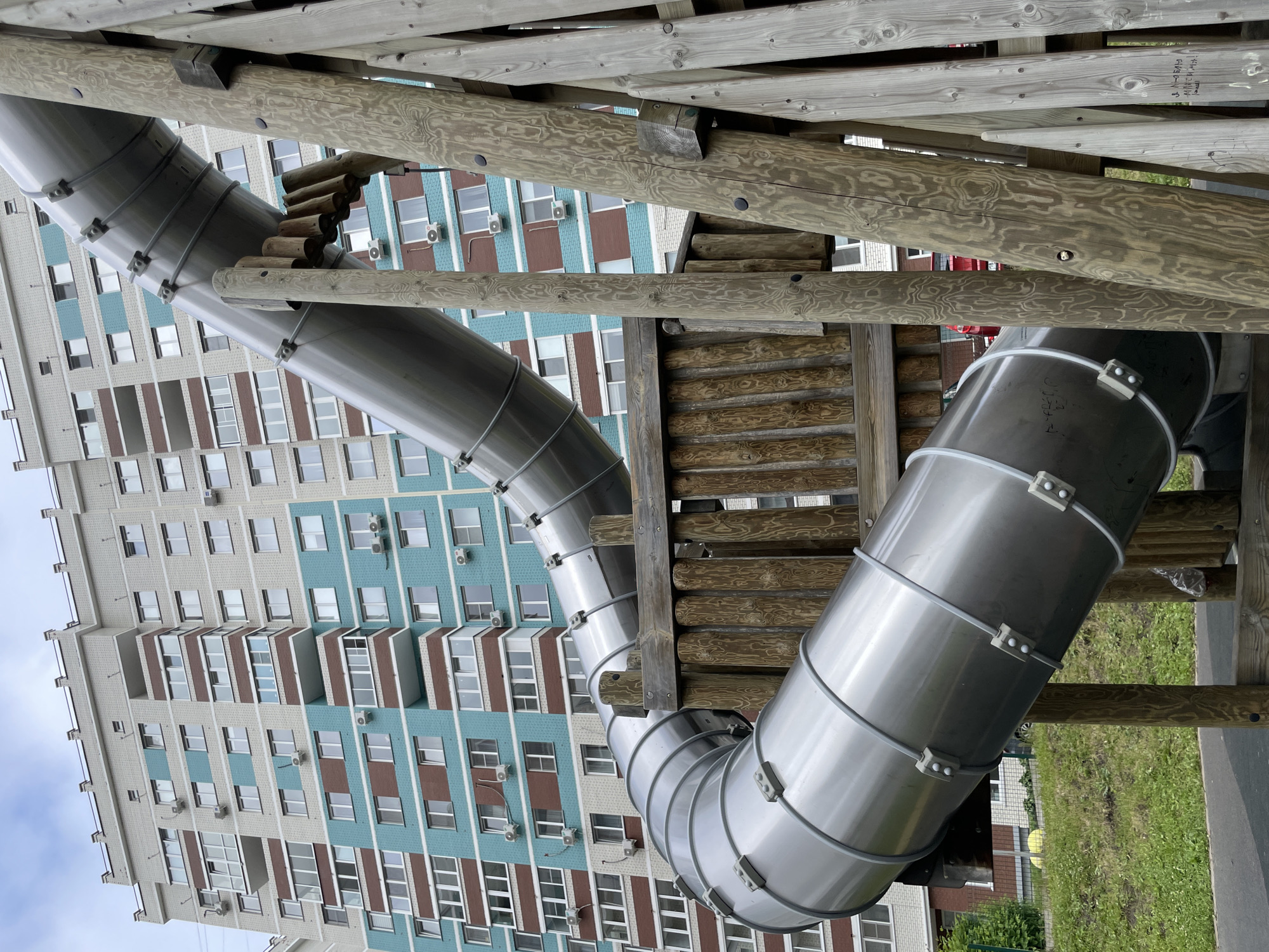 The image, rotated counter-clockwise, depicts a metal tube slide and a wooden playground structure against the backdrop of a large apartment building. The scene is outdoors, likely a playground area adjacent to a residential complex in Izhevsk, Russia.
The prominent feature is a curvilinear, silver-colored metal tube slide, constructed from multiple bolted sections. The slide is supported by a robust wooden structure made of light-colored logs and planks, characteristic of a playground. The wood shows a distinct grain pattern and some wear. No people are visible, so no specific activity is taking place, but the presence of the slide and structure implies a recreational setting.
In the background, a multi-story apartment building dominates the view. Its facade features a repeating pattern of light grey, light blue, and brown panels, suggesting a block apartment style. Numerous windows are visible, many with external air conditioning units attached. The sky, partially visible at the upper left (when oriented correctly), appears overcast.
Notable details include handwritten text on one of the wooden planks in the playground structure, which is difficult to decipher fully but appears to include "3-й этаж" (3rd floor) and other Cyrillic script. Faint alphanumeric markings, possibly production codes like "7-02-01", are also visible on the metal slide sections. The ground below the playground structure is covered in green grass, with some paved areas and shrubbery visible towards the bottom right. The image is taken during the daytime, with soft, diffused lighting.