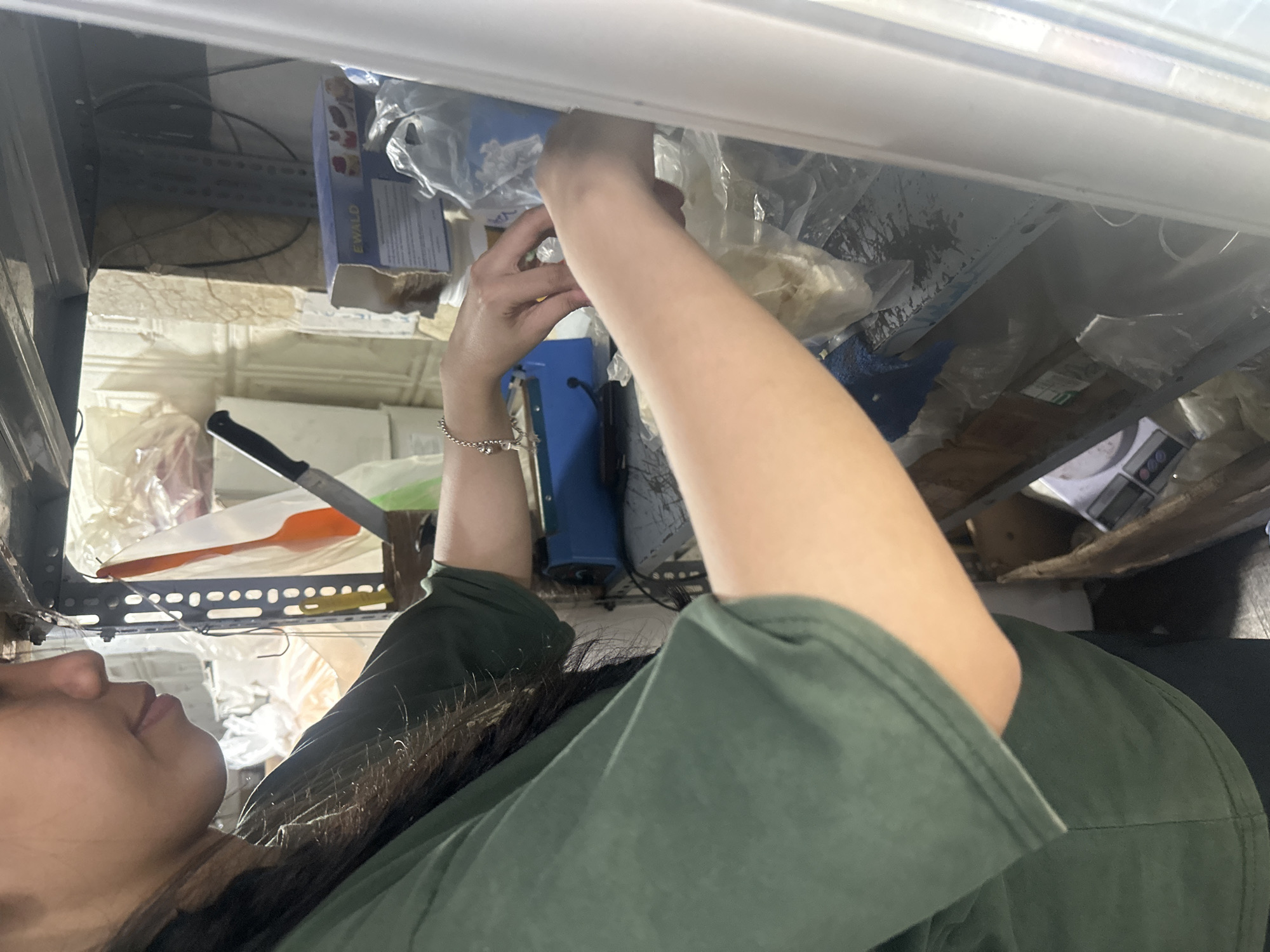 A person is reaching into a cluttered shelf space. The person has brown hair and is wearing a green shirt and a silver bracelet. Visible objects on the shelves include a knife, an orange spatula, boxes, papers, and plastic bags containing various items. The location is likely a storage area in Nha Trang Ward, Vietnam, based on other data available.