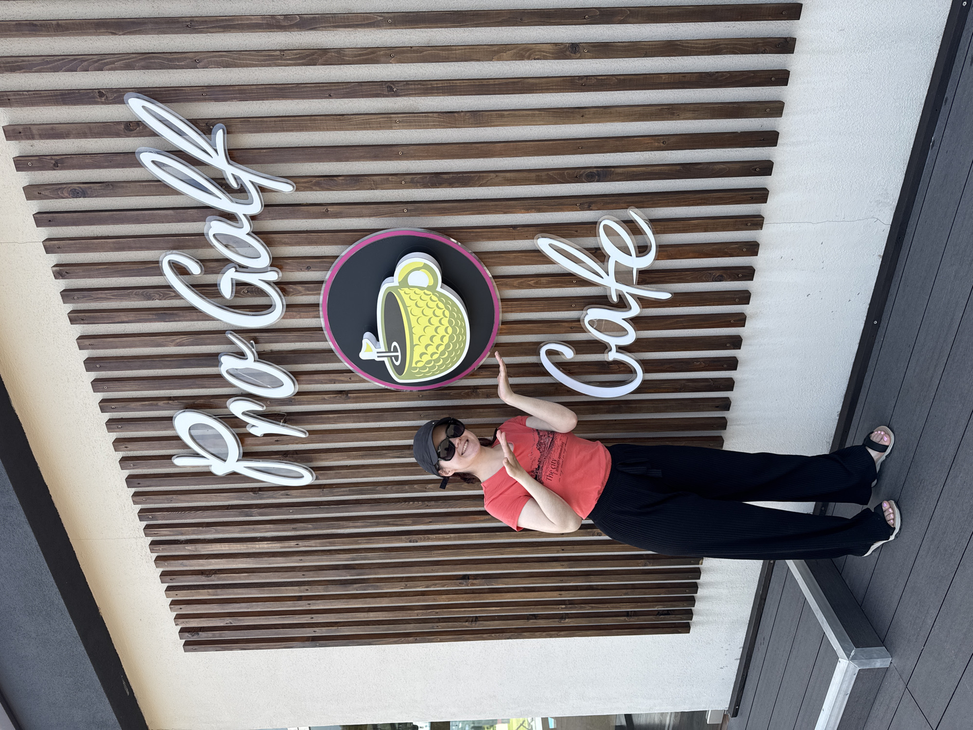 A woman stands beneath a sign that reads "Pro Golf Cafe." The sign is mounted on a structure with horizontal wooden slats. A circular logo featuring a golf ball inside of a cup is situated between the words "Pro Golf" and "Cafe." The woman wears a red t-shirt, black pants, sandals, sunglasses, and a baseball cap. The cafe is located in the city of Sirius, Russia.