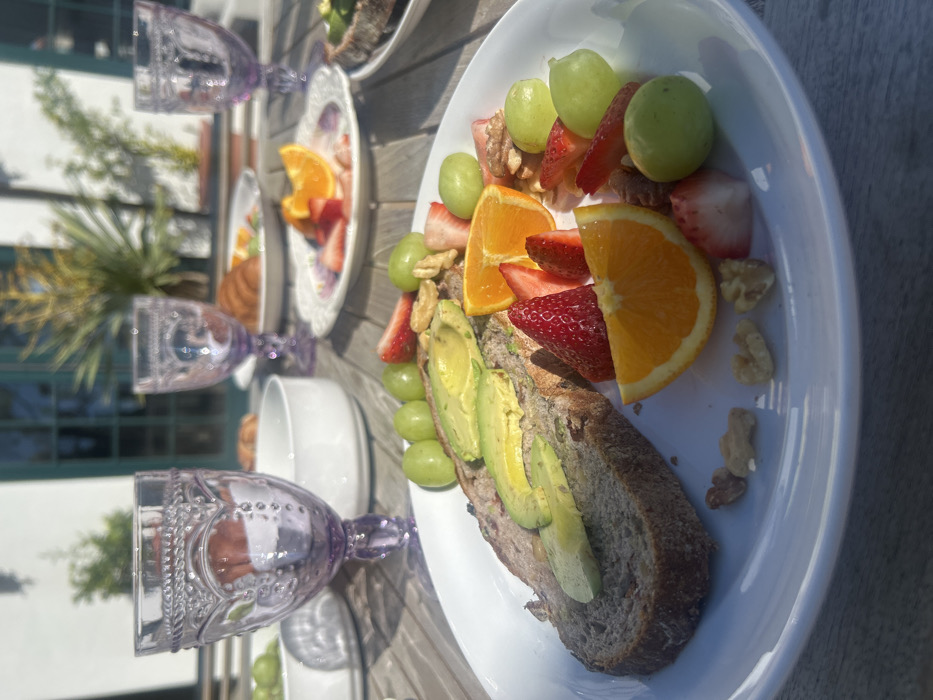 The image captures a vibrant outdoor breakfast or brunch scene set on a sunny day. The main focus is on a white oval plate filled with an appealing meal.
On this prominent plate, there is a thick slice of dark, whole-grain bread, generously topped with slices of ripe green avocado. Accompanying the avocado toast is a colorful assortment of fresh fruit, including bright green grapes, red strawberries (some whole, some sliced), and wedges of orange. Scattered among the fruit are pieces of walnuts and what appears to be some granola or cereal, adding texture to the dish.
In the background, the scene reveals more of the table setting. Several ornate, stemmed glasses, made of what appears to be purple or lavender-tinted pressed glass with intricate patterns, are placed on a light-colored wooden table. Another white plate with a decorative, possibly floral, rim holds more fruit, including orange slices and strawberries. A white bowl is also partially visible, suggesting other elements of the meal.
The setting appears to be outdoors, likely on a patio or deck, as indicated by the wooden table and the lush green foliage and potted plants seen in the blurred background. A building with green trim or window frames is also visible. The bright sunlight creates strong highlights on the food and tableware, casting soft shadows and contributing to a fresh and inviting atmosphere. The overall impression is one of a leisurely and wholesome meal enjoyed in pleasant, warm weather. There is no visible text in the image.