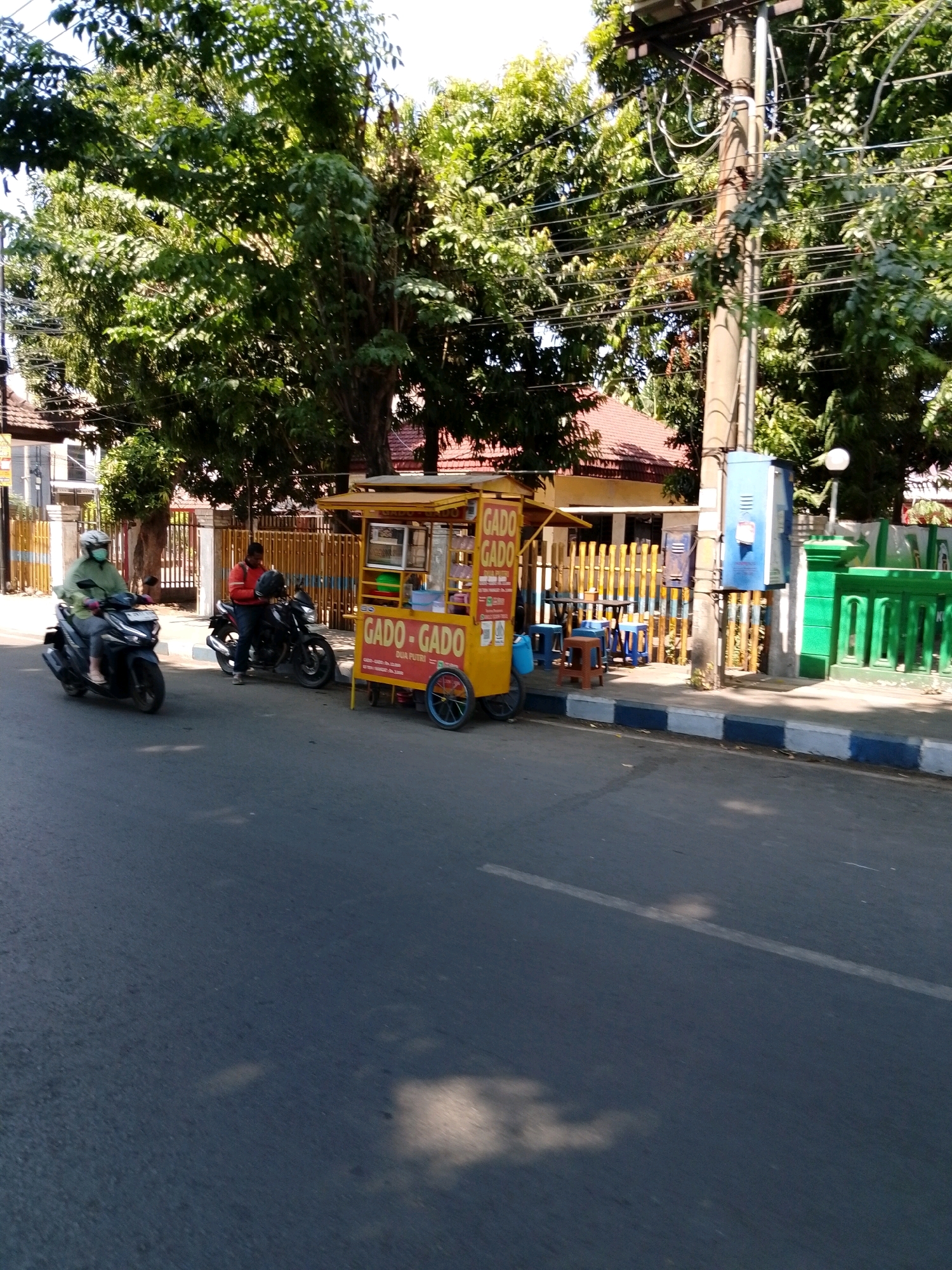 A sunny day scene on a street in Pasuruan, Indonesia. A person on a scooter with a green jacket and white helmet is riding down the asphalt road. Behind them, another person sits on a motorcycle. To the right, a yellow food cart labeled "GADO-GADO" is parked. The cart has a blue wheeled base and a small display window. In the background, a low wooden fence separates the sidewalk from a building with a red roof. A utility pole with electrical wires and a blue electrical box stands near the food cart. Green metal railings are visible to the right of the utility pole. Trees with lush green foliage line the area. The road has a white line marking and shadows from the trees are cast on the asphalt.