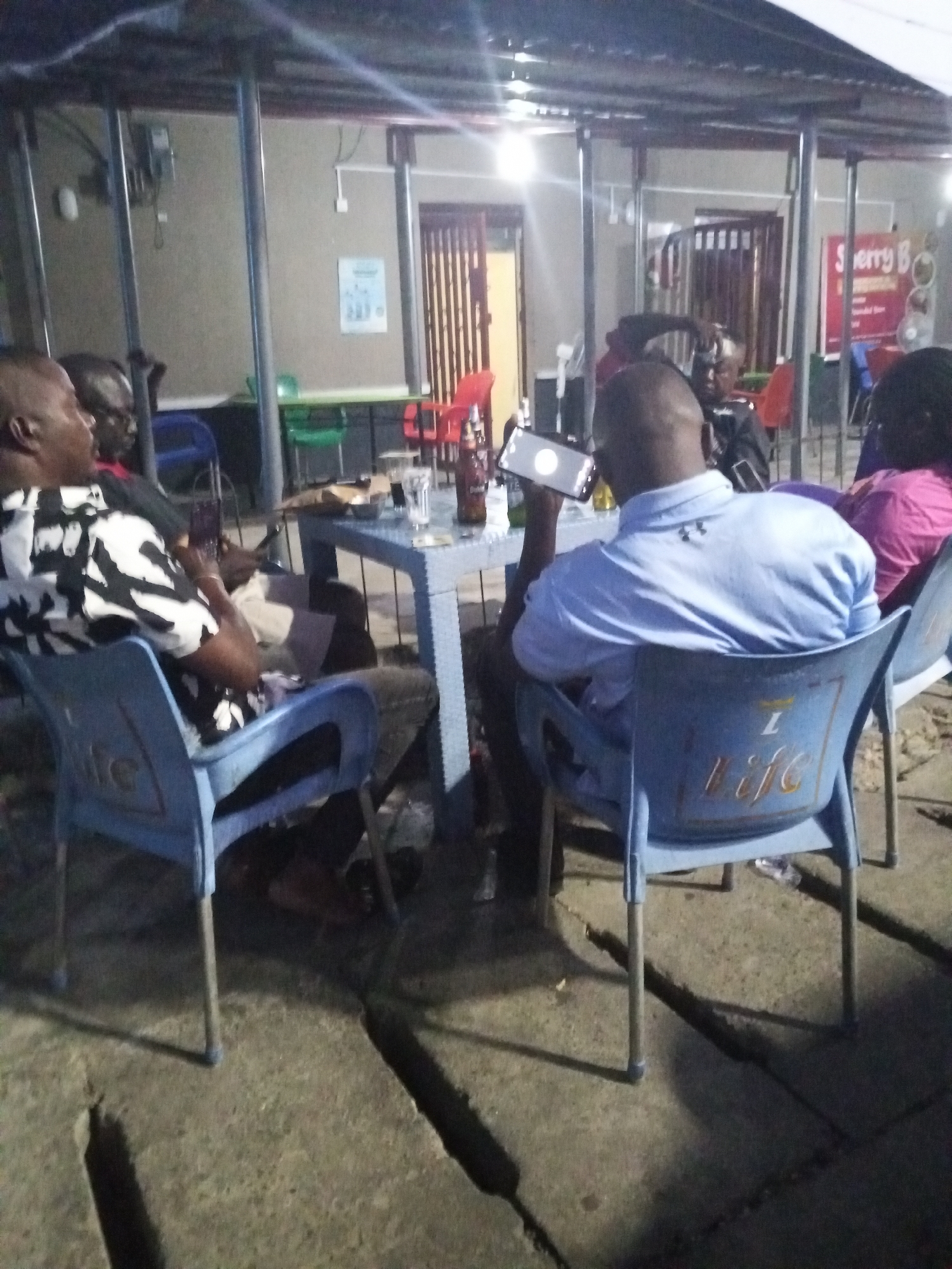 Four men and a woman are gathered around a blue plastic table at an outdoor bar or restaurant. The bar appears to be named "Sherry B." The men are of African descent and are dressed in casual attire. One man is holding a smartphone in front of him. There are drinks on the table and plastic chairs surrounding it. The location is Lokoja, Nigeria.