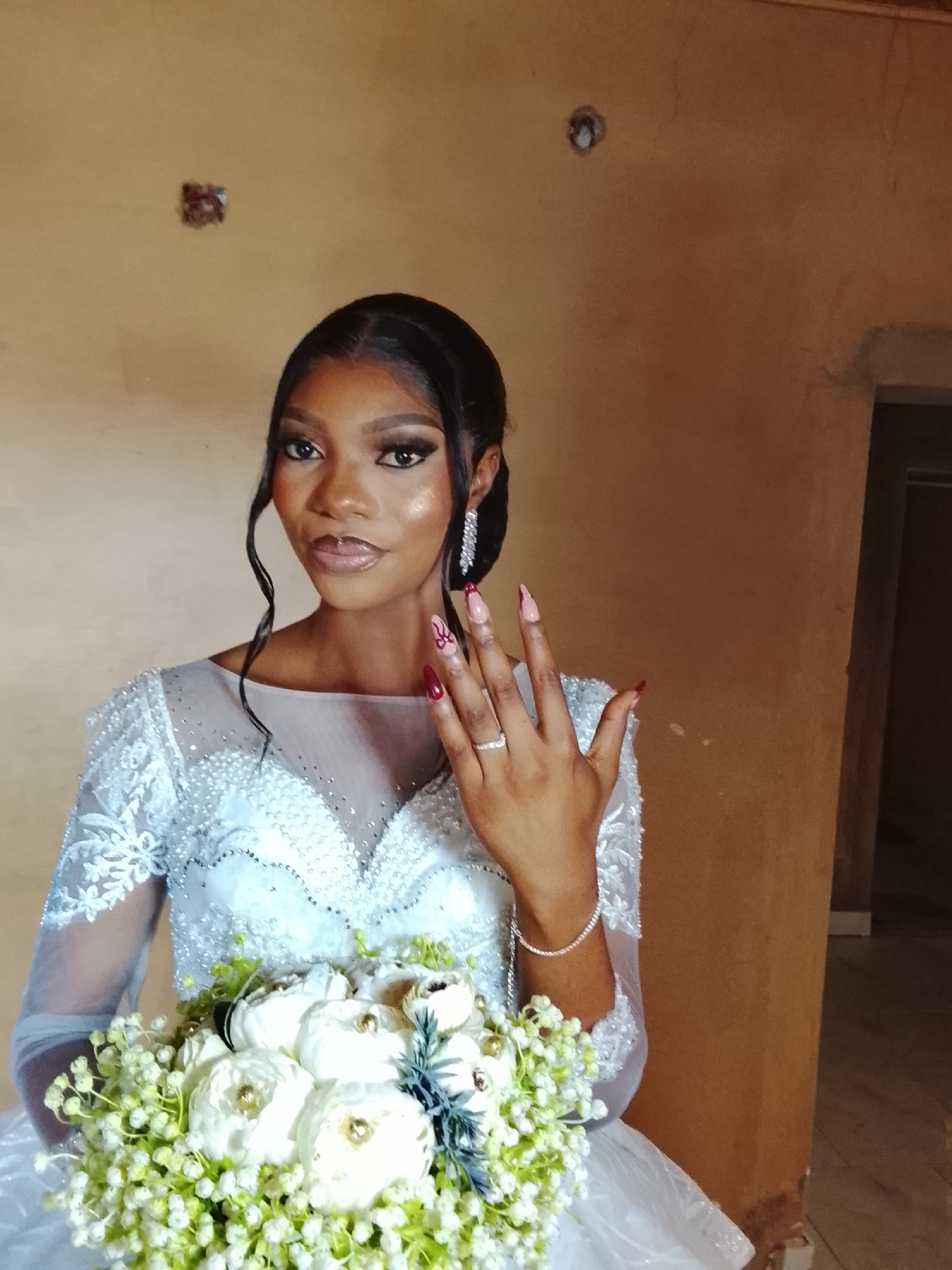 A young woman, likely a bride, is captured in a close-up, half-body portrait indoors, presumably in Jos, Nigeria. She stands gracefully, holding a lush white and light-green floral bouquet, and extends her left hand slightly to showcase a ring on her ring finger.
The woman has dark skin, an oval face, and a warm, pleasant expression with a subtle smile. Her dark hair is styled in an elegant updo, with soft, curly tendrils framing her face. She wears full, glamorous makeup, featuring defined eyebrows, dramatic winged eyeliner, false eyelashes, and glossy, peachy-nude lips. Her long, pointed nails are artfully manicured, some with deep red or maroon tips, and others displaying intricate red designs over a nude base.
She is dressed in a beautiful white wedding gown, which features an illusion neckline and long sleeves made of sheer fabric adorned with delicate lace patterns and sparkling bead or sequin embellishments. The bodice also features intricate beading and a V-neck design under the sheer overlay. She accessorizes with prominent, sparkling drop earrings and a delicate diamond-like bracelet on her right wrist.
The background is a plain, warm-toned wall, appearing light brown or ochre, showing some slight imperfections and a couple of indistinct dark marks higher up. To the right, part of a darker brown wall or doorway is visible, suggesting a simple indoor setting. The lighting is soft, highlighting her features and the details of her attire and accessories.