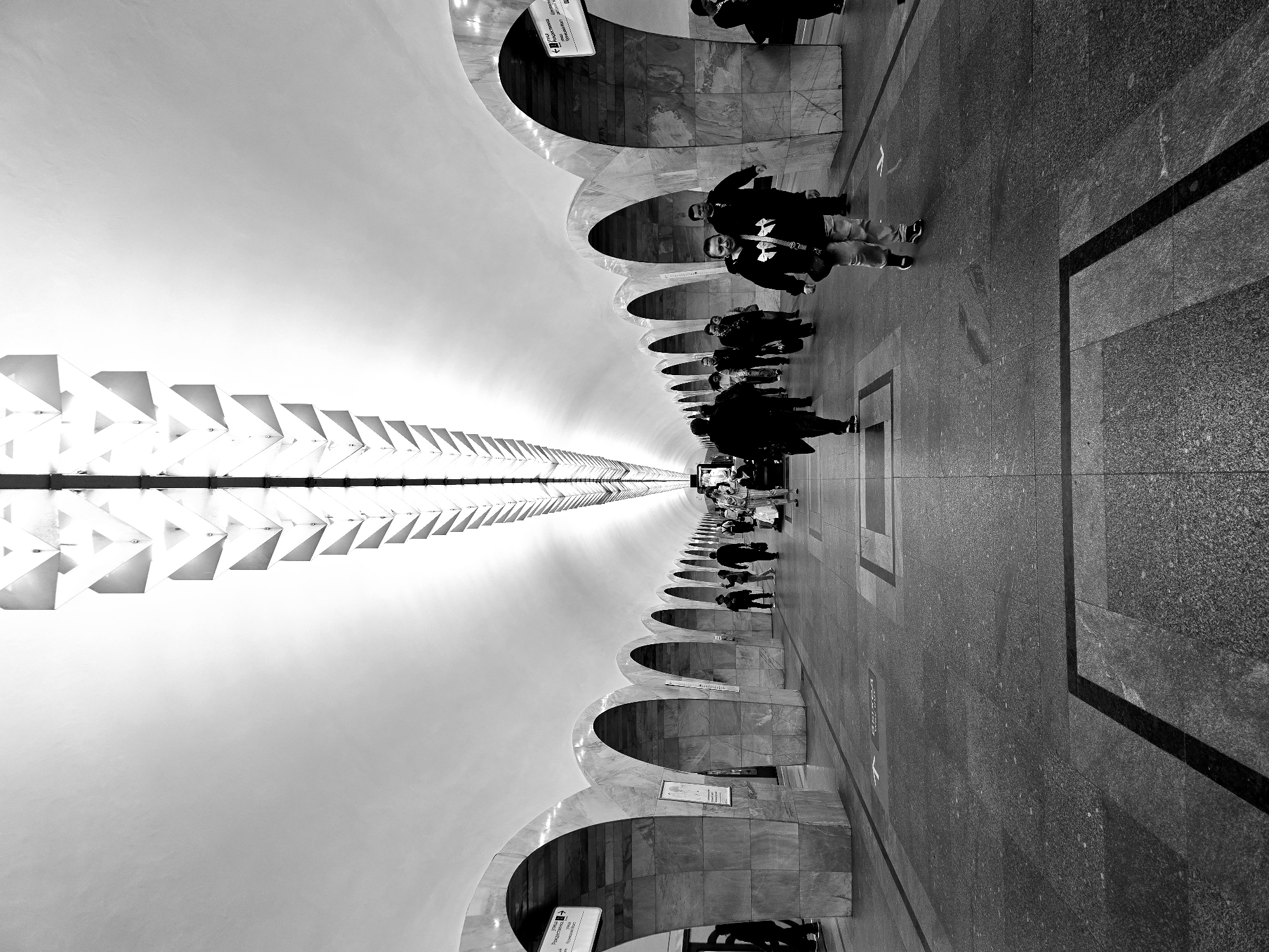 This black and white, horizontally oriented photograph depicts a subway station in Moscow, Russia. The image, taken from a ground-level perspective, shows a tunnel with an arched roof and a series of arched portals along one side. The tunnel appears to stretch into the distance, creating a sense of depth. People are seen walking along the tunnel, with several appearing closer to the viewer and becoming smaller as they recede into the distance. A lighting fixture is suspended from the center of the tunnel roof, casting light onto the surrounding area. The floor is made of stone. A black border of a square is present on the right side of the image. The overall environment suggests a busy, urban setting within Moscow's subway system.