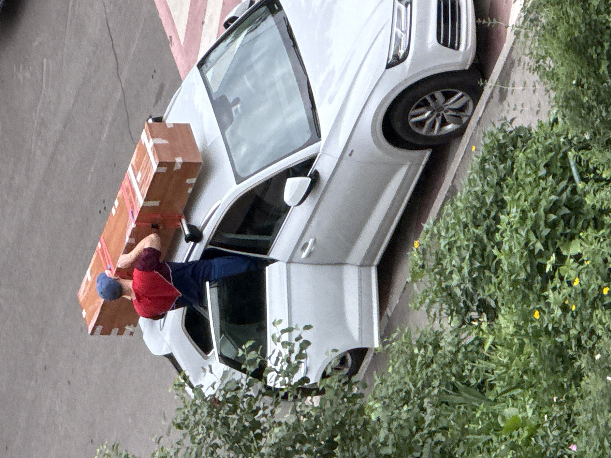 A person wearing a red and blue shirt and a blue cap is standing on the roof of a silver car, lifting a large, wooden object that is wrapped in tape. The car's driver's side door is open. The scene appears to be on a street in Khimki, Russia, with trees and bushes in the foreground.